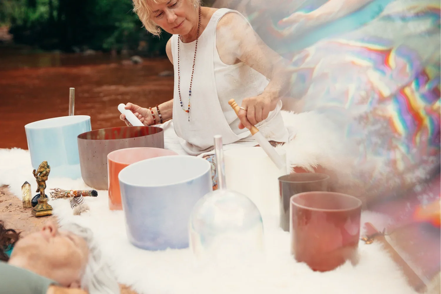 A woman playing crystal singing bowls outdoors by a river, with various small spiritual and decorative items on the table.