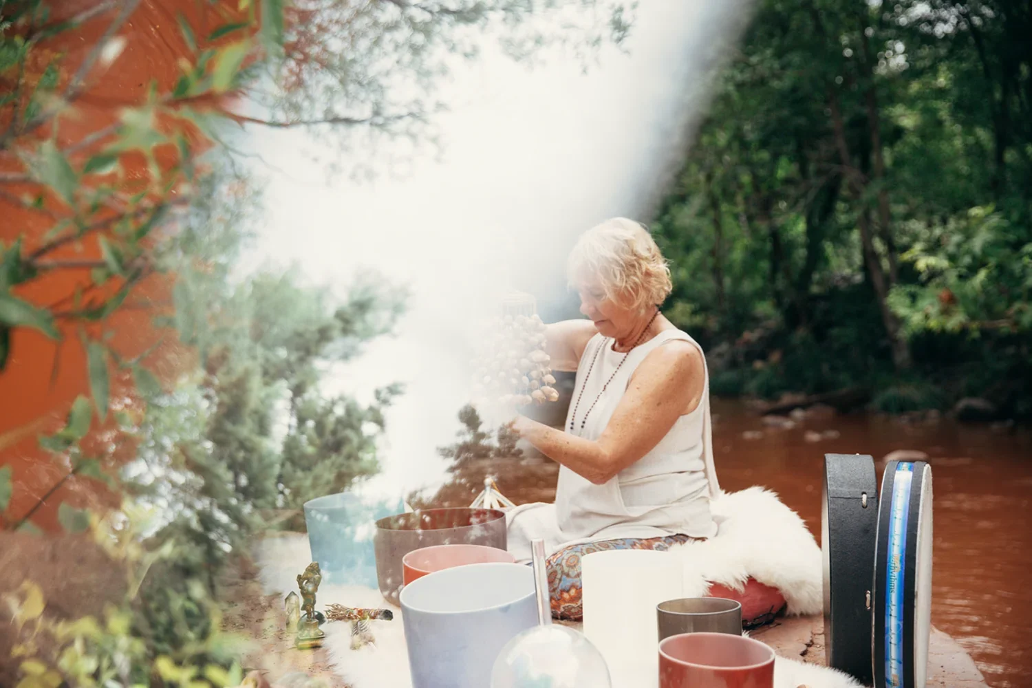 An elderly woman with white hair, dressed in white, sitting outdoors near a river or stream, surrounded by greenery, and holding a string of beads or mala.