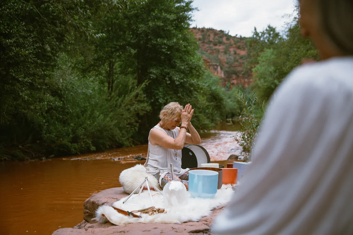 A woman sitting cross-legged on a rock in a river, practicing yoga with hands in prayer position, outdoors surrounded by trees and mountains, with various singing bowls and a drum nearby.