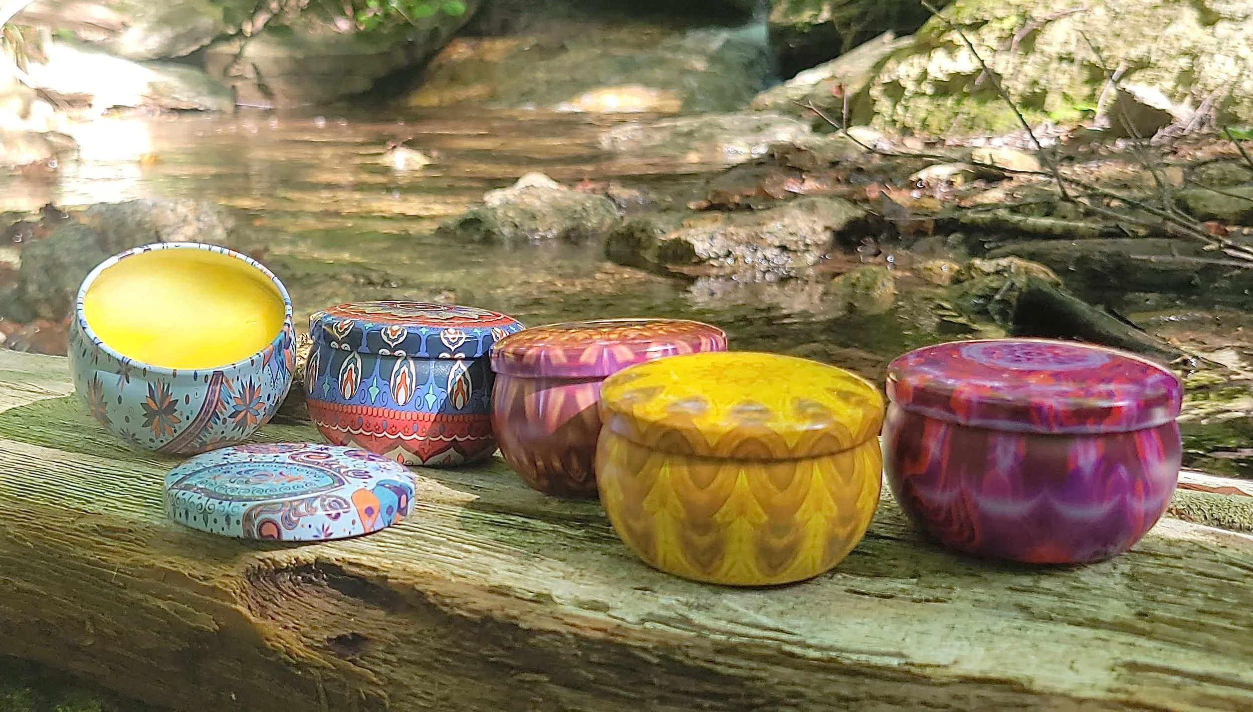 Colorful tins of Mind Your Own Beeswax hand balm arranged outdoors in a mountain setting.