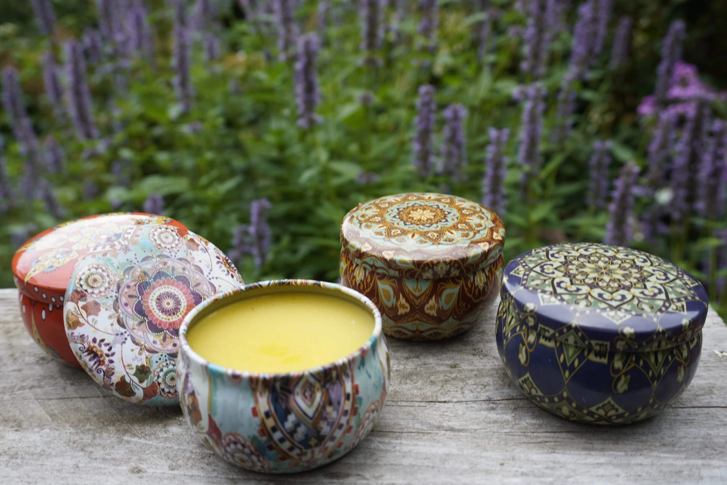 Decorative tins of Mind Your Own Beeswax hand balm displayed outdoors, with one open tin showing the golden balm.