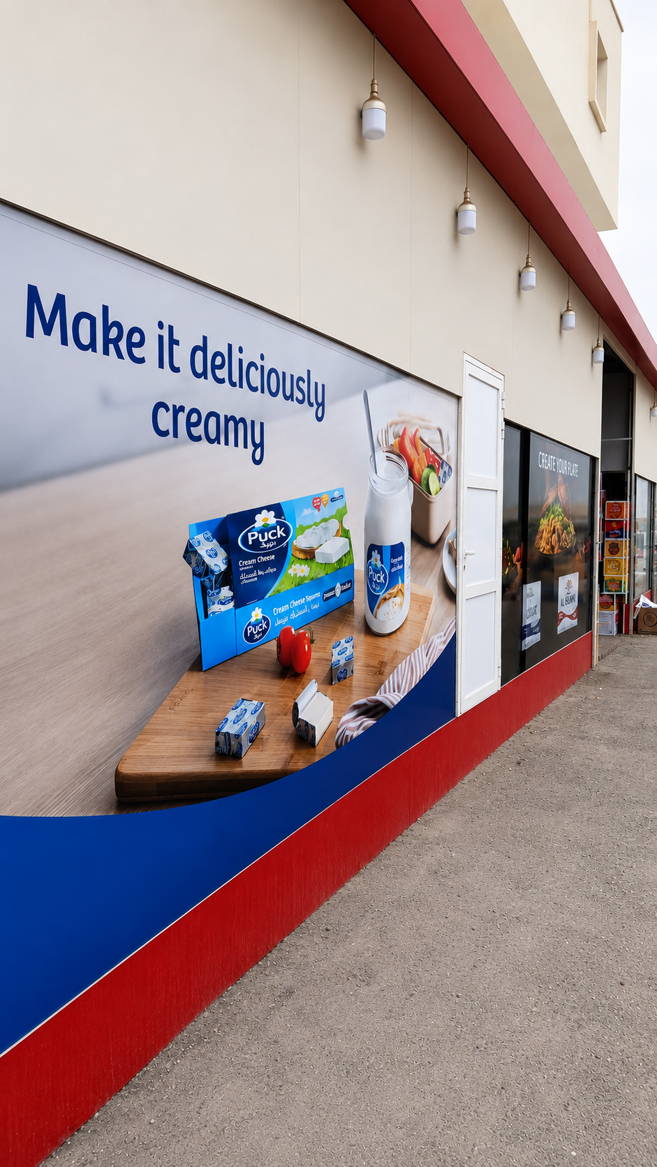 Store window display promoting creamy dairy products with a large sign that says 'Make it deliciously creamy.' The display includes cartons of Puck cream cheese, a bottle of milk with a Puck label, and various dairy products on a wooden surface.