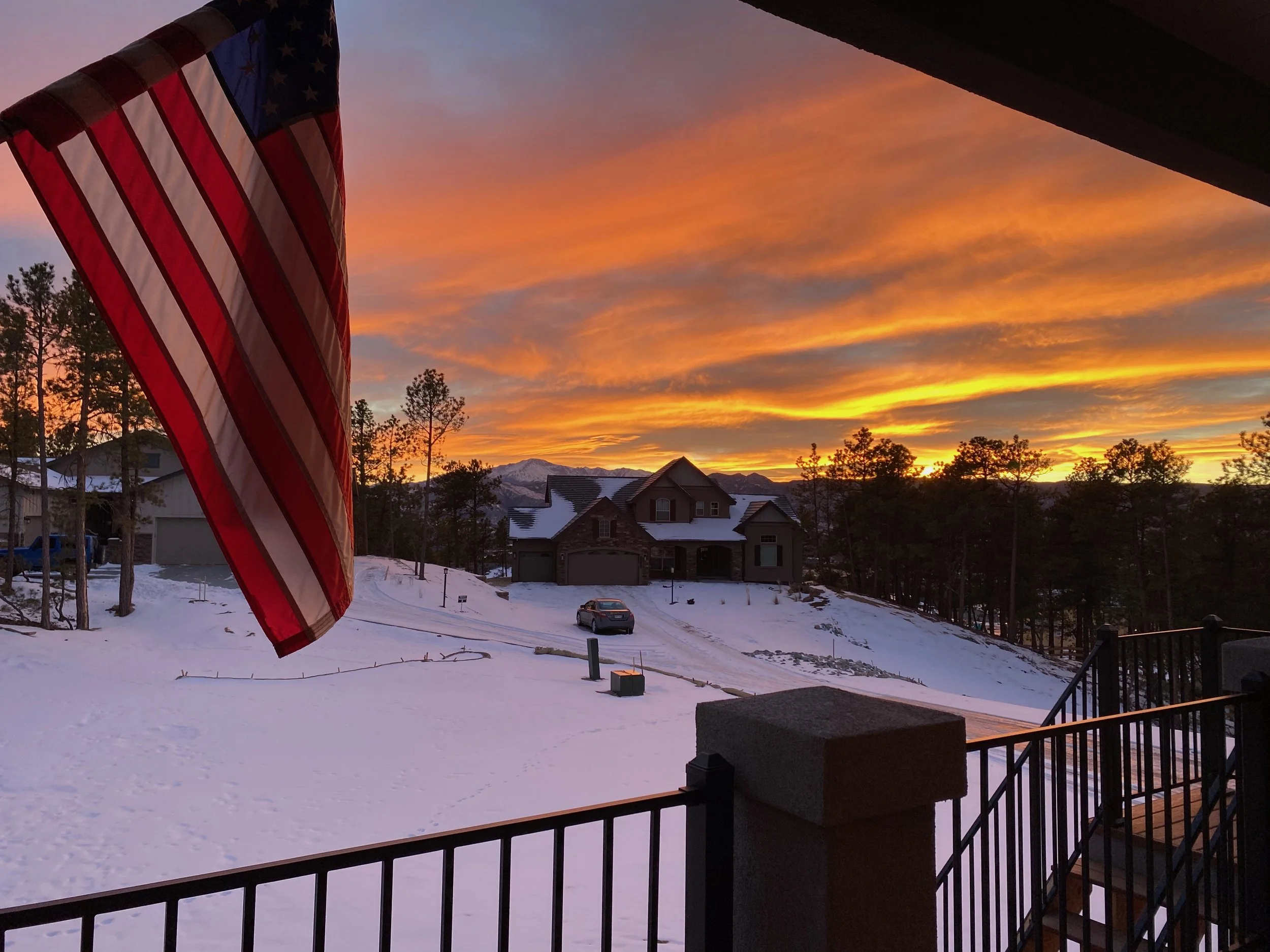 A front yard covered in snow with houses, trees, and a mountain in the background during sunset. An American flag is hanging on a pole on the left side of the image.