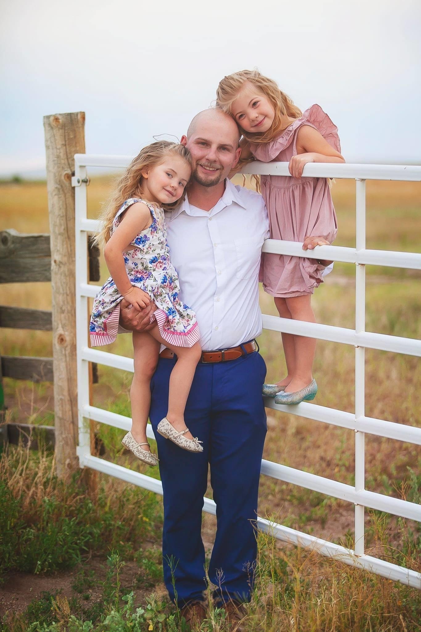 Man with two young girls outdoors by a white gate, smiling and leaning on each other, in a rural setting with grass and a wooden fence.