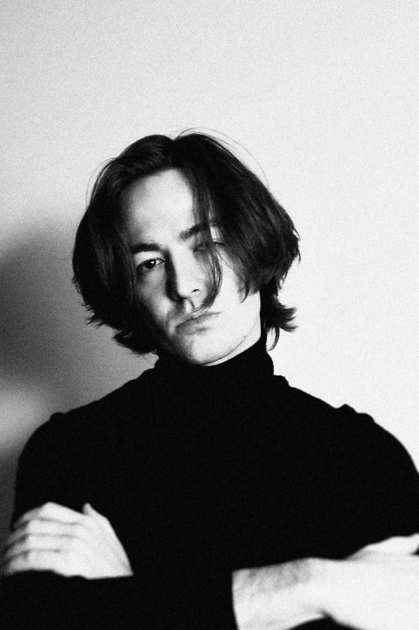 A black and white portrait of a person with shoulder-length hair, wearing a turtleneck sweater, looking at the camera with a neutral expression.
