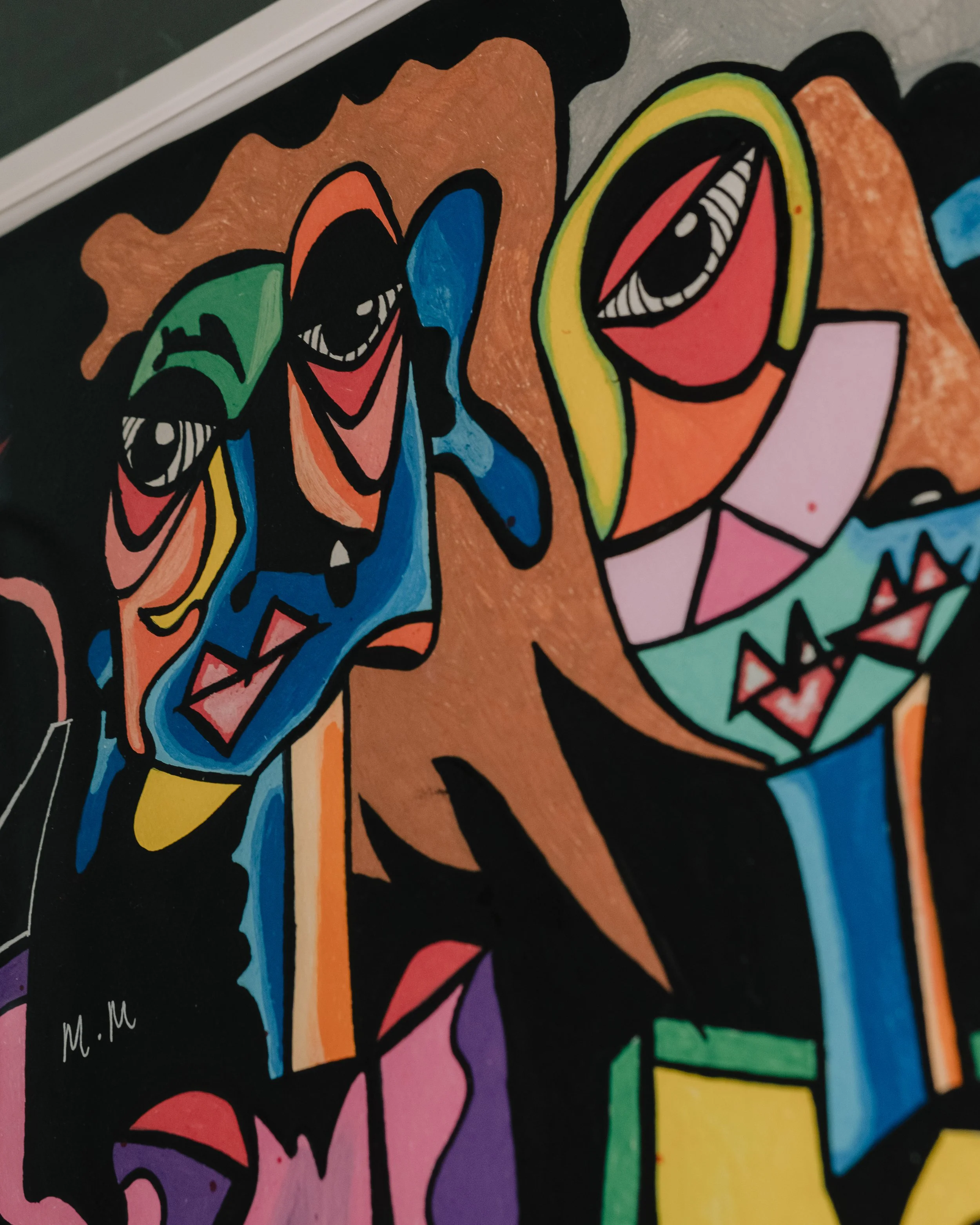 Close-up of a colorful graffiti or mural with stylized faces, eyes, and abstract shapes in vibrant colors including orange, blue, green, yellow, pink, and purple, set against a black background.