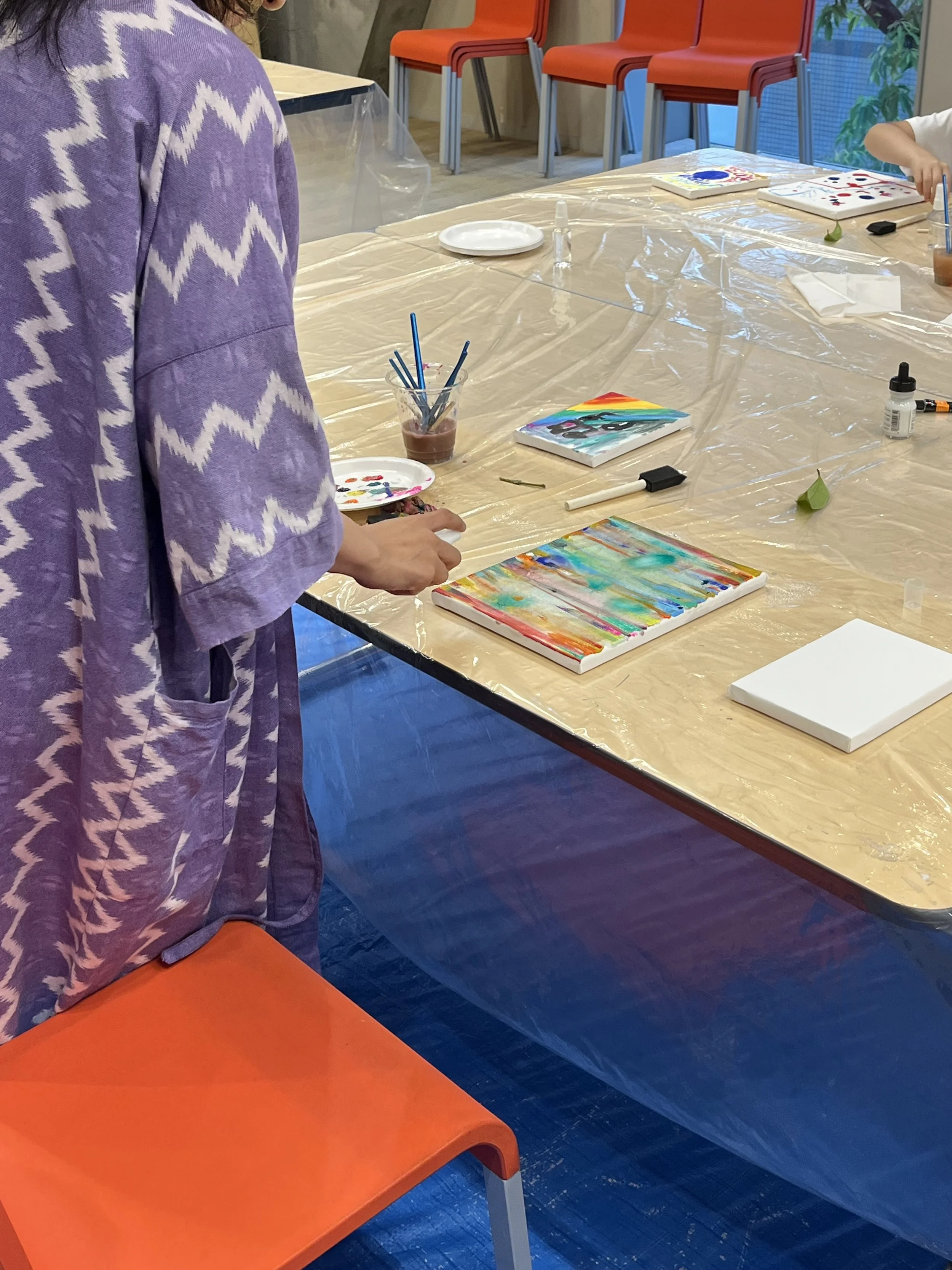 A person with a purple and white zigzag patterned shirt or jacket standing next to an orange chair at a table covered with a clear plastic sheet. On the table are a white plate, a cup with paintbrushes, small bottles, and canvas paintings with colorful abstract designs. In the background are stacked orange chairs against a window showing outdoor greenery.