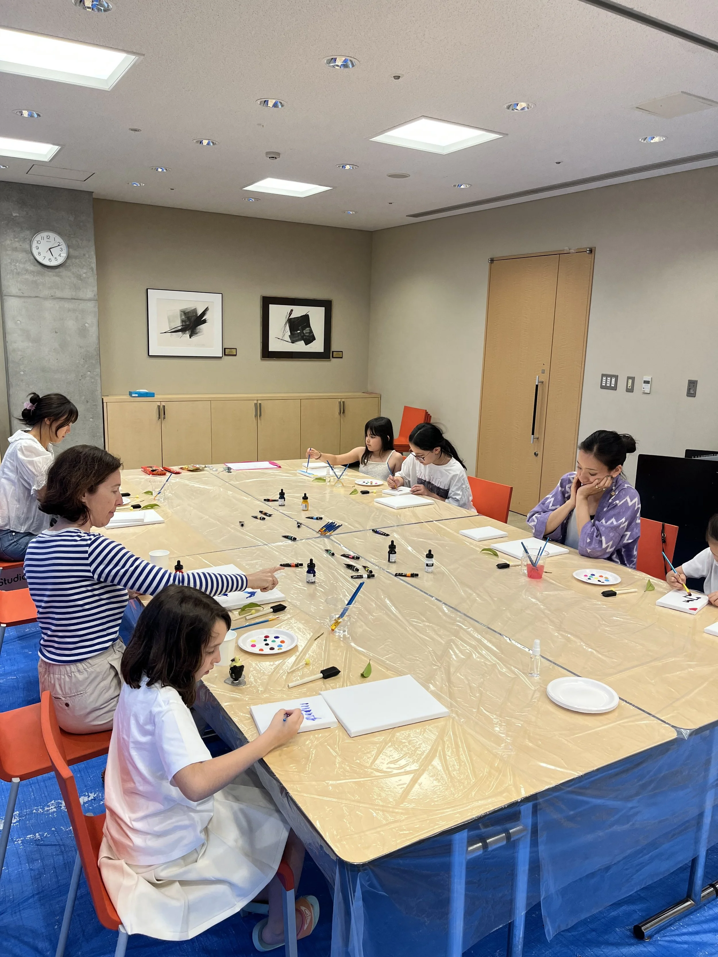 Children and adults sit around a large table covered with a plastic sheet, painting with brushes and watercolor paints in a well-lit room. There are art supplies scattered on the table, and framed abstract artwork hangs on the beige wall behind them.
