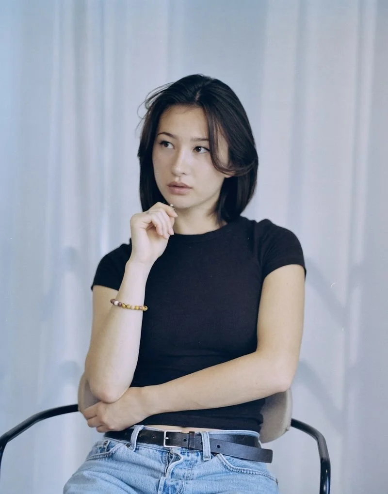 A young woman with shoulder-length dark hair, wearing a black t-shirt and light jeans, sitting on a chair with her left arm crossed and her right hand touching her chin, looking thoughtfully to her left in front of a white curtain background.