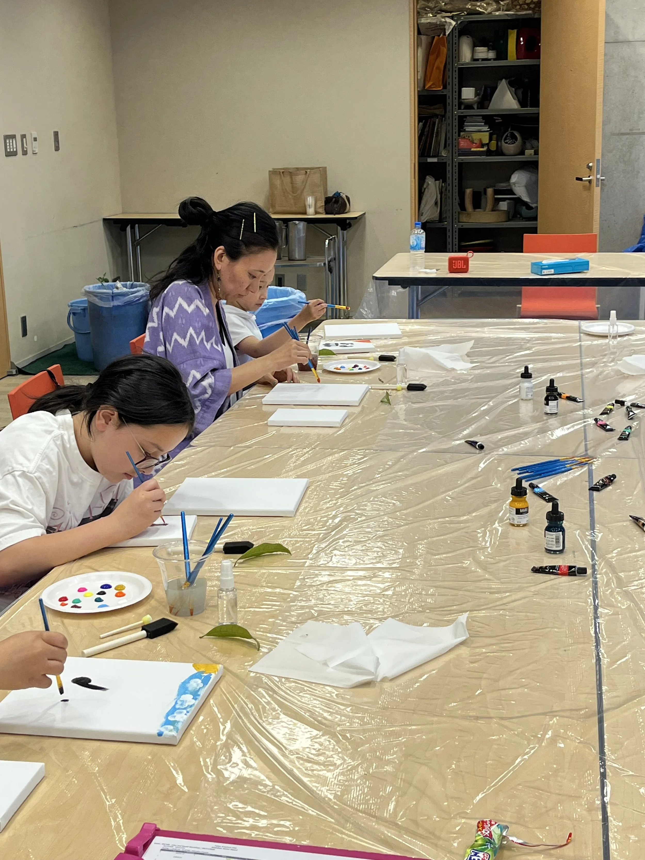 People sitting at a long table covered with a plastic sheet, engaged in painting on canvases with paints and brushes, in an indoor setting.