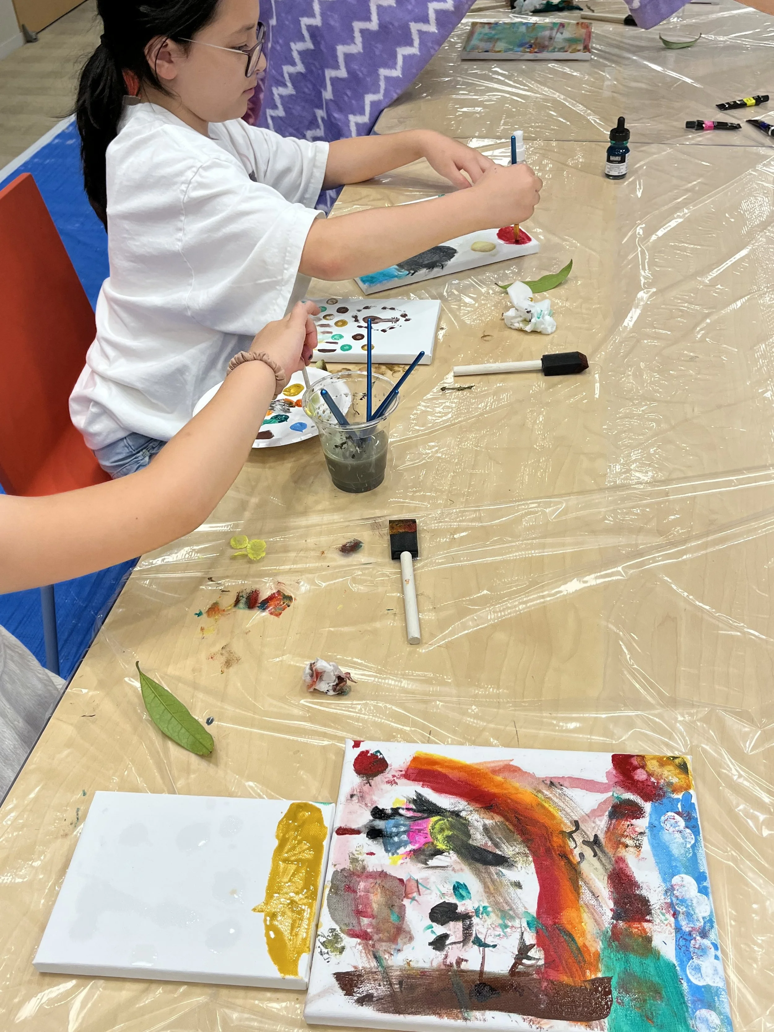 A young girl with glasses and a white shirt is painting on a canvas using bright colors. The table is covered with a clear plastic sheet, and there are various art supplies around, including paint palettes, brushes, and a small bottle of paint. The girl is focused on creating her artwork, which features abstract and colorful patterns.