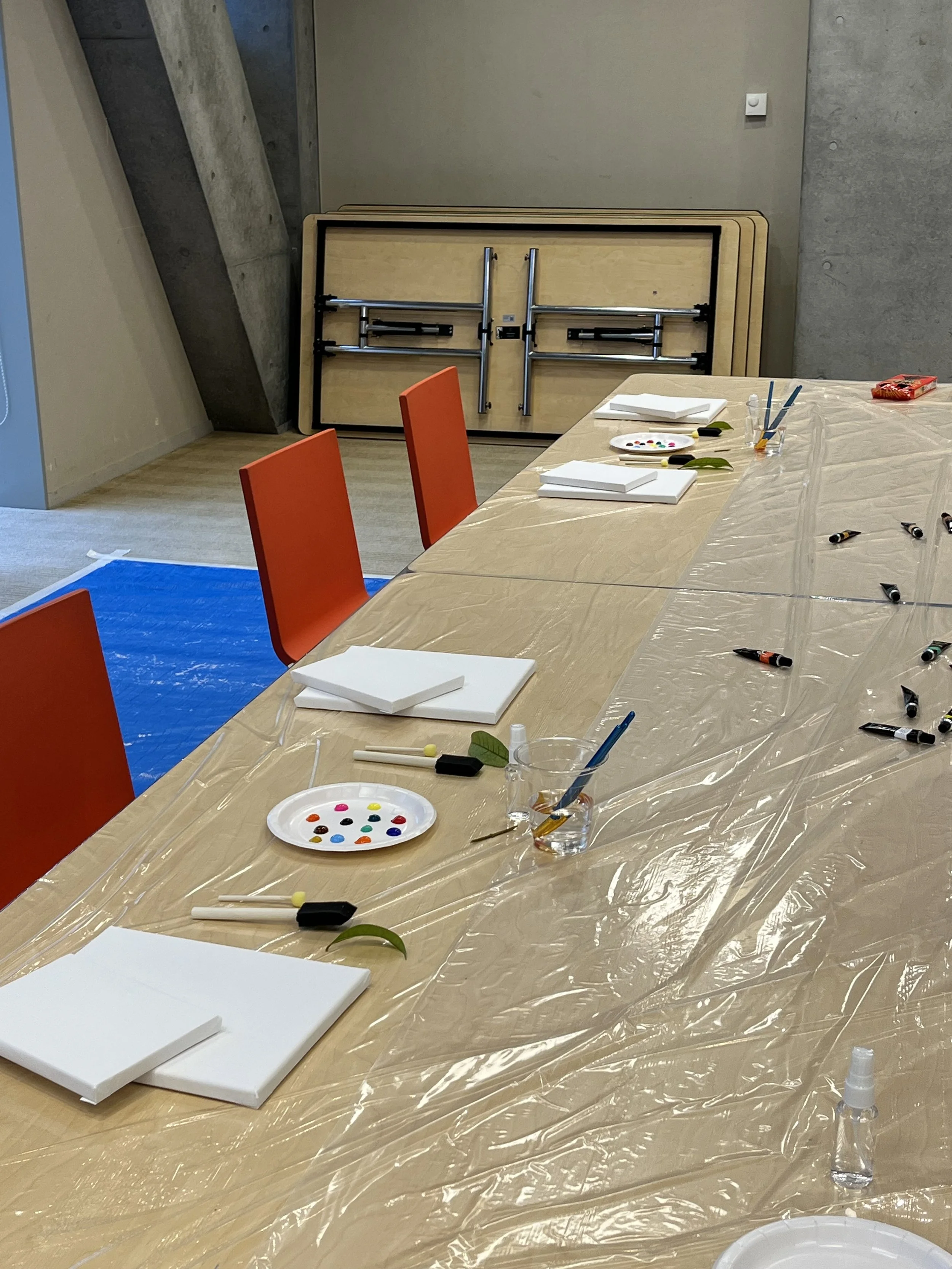 A room with a long wooden table covered with a plastic sheet, surrounded by orange chairs. On the table, there are white canvases, paint palettes, brushes, and paint bottles. A stack of three folding tables leans against the wall in the background, and the room has concrete walls and floors.