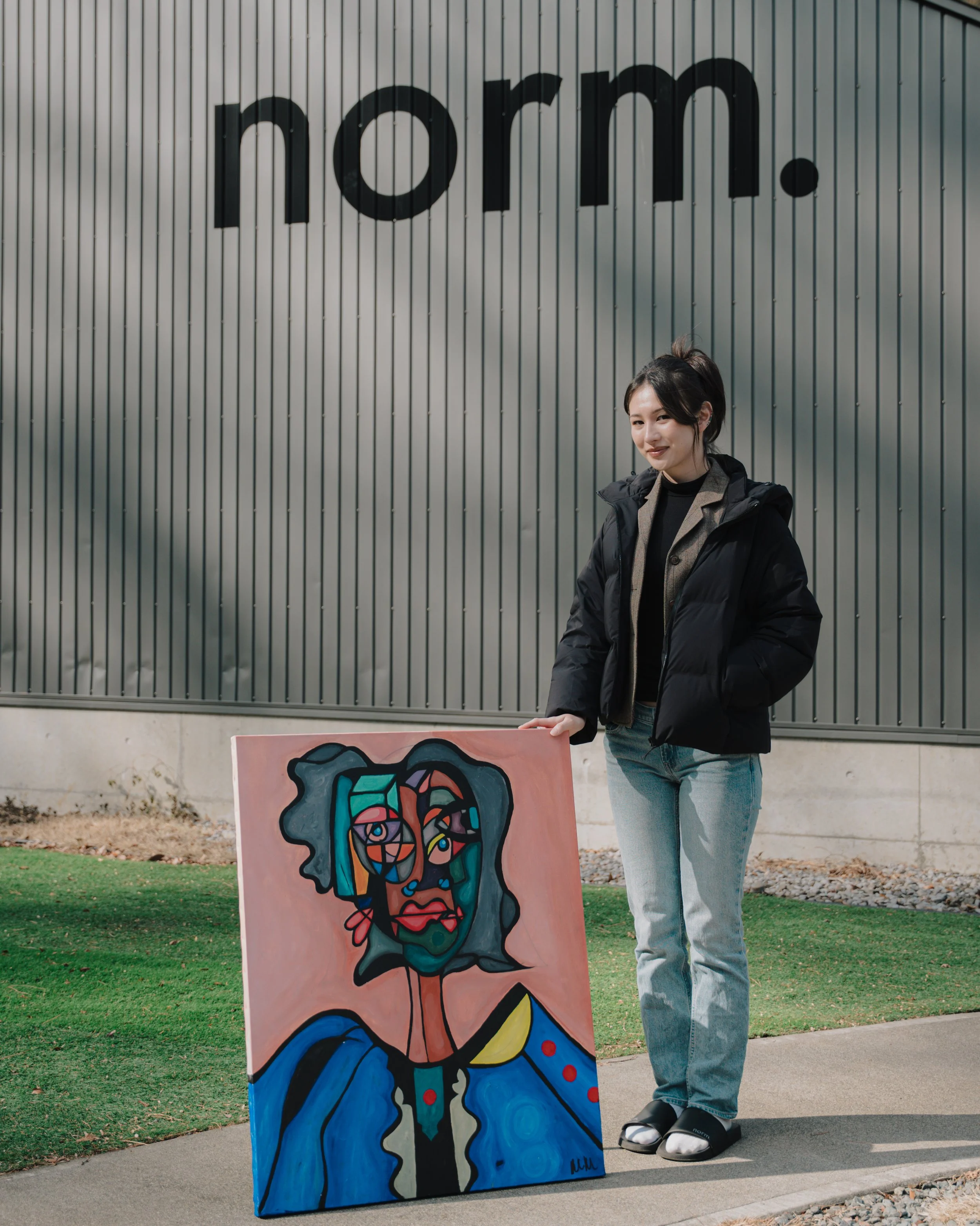 A young woman standing outdoors next to a colorful abstract painting of a face, on a sidewalk with grass and a building in the background.