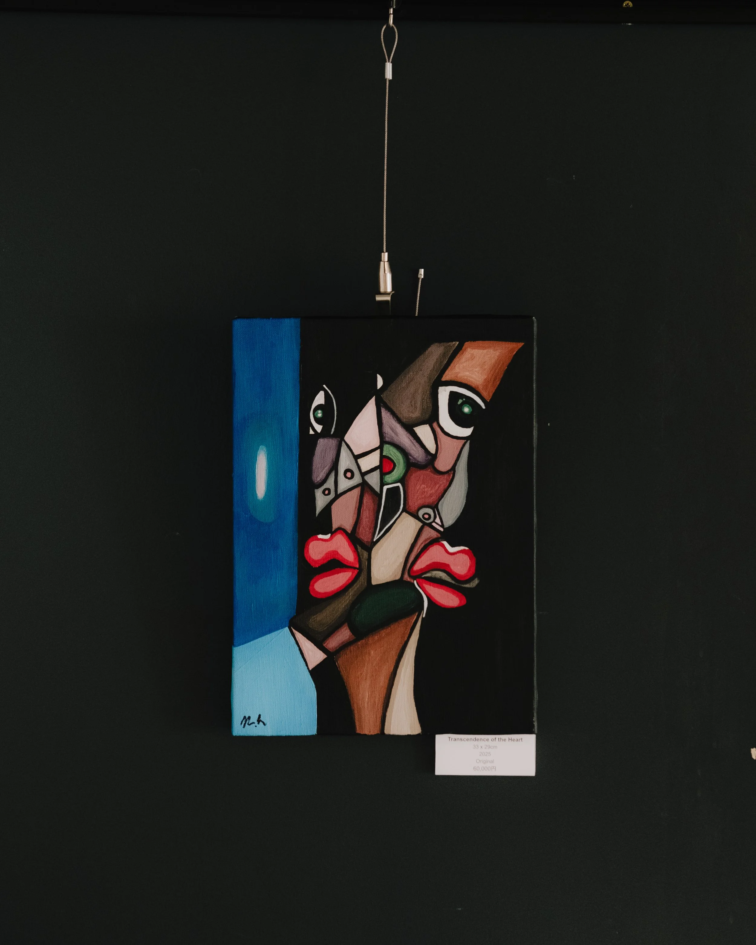A colorful abstract painting titled "Transcendence of the Heart" featuring geometric shapes of human faces, hearts, and various colors like blue, pink, green, and brown, hanging on a black background.