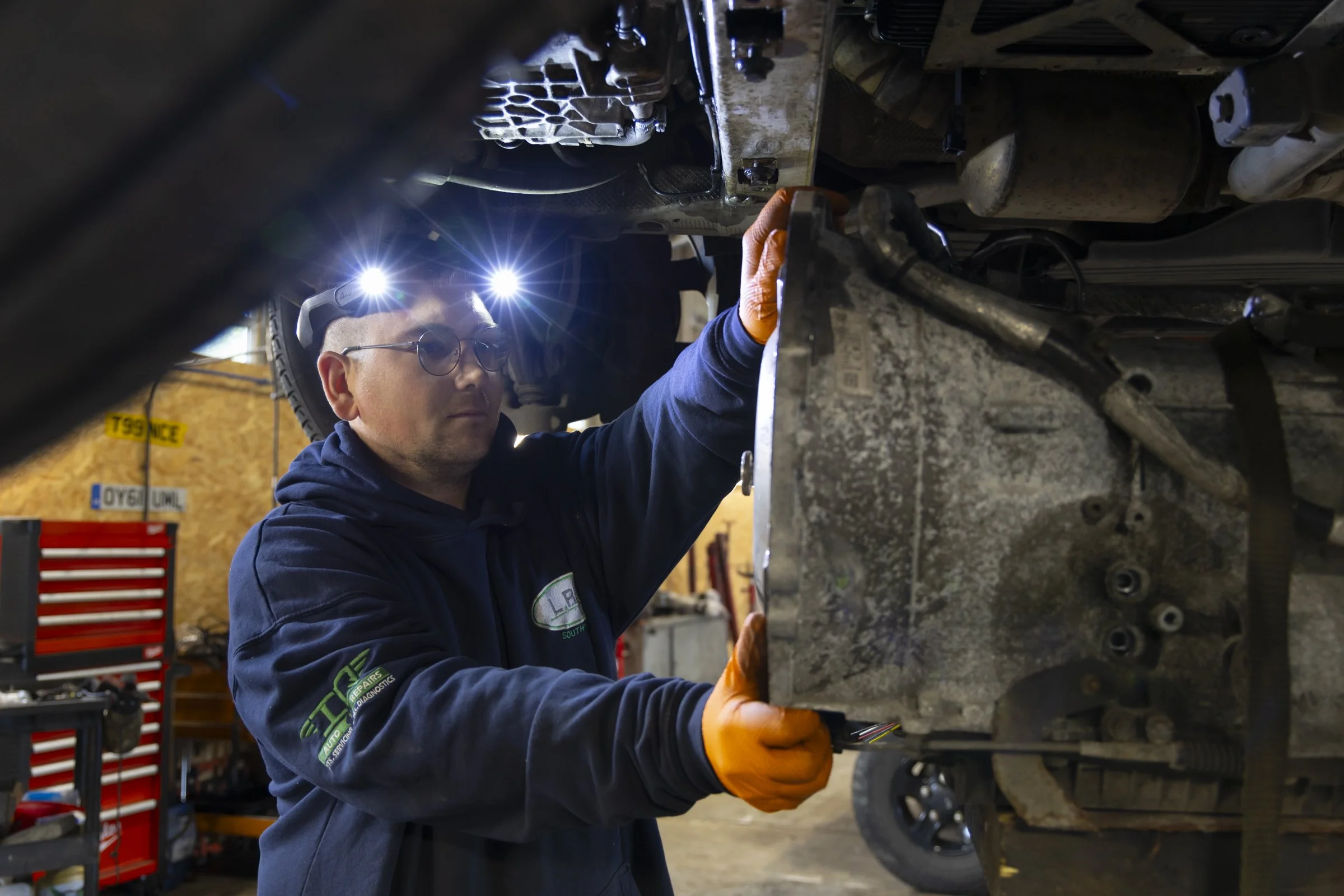 A mechanic wearing safety glasses and orange gloves inspects a large, dirty vehicle transmission in a garage.