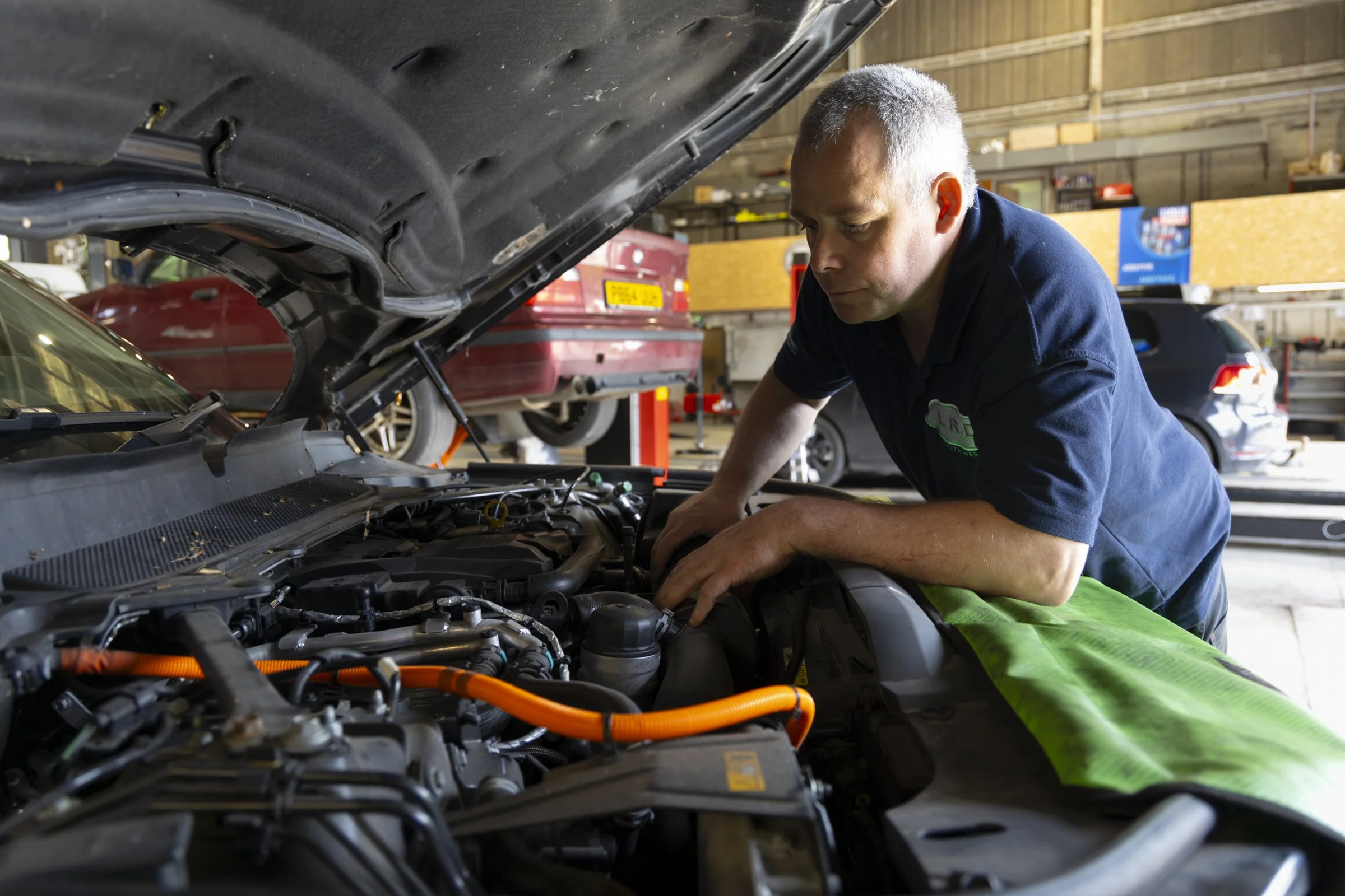 A mechanic with gray hair working under the hood of a car inside a garage surrounded by other vehicles and shelves with tools and car parts.