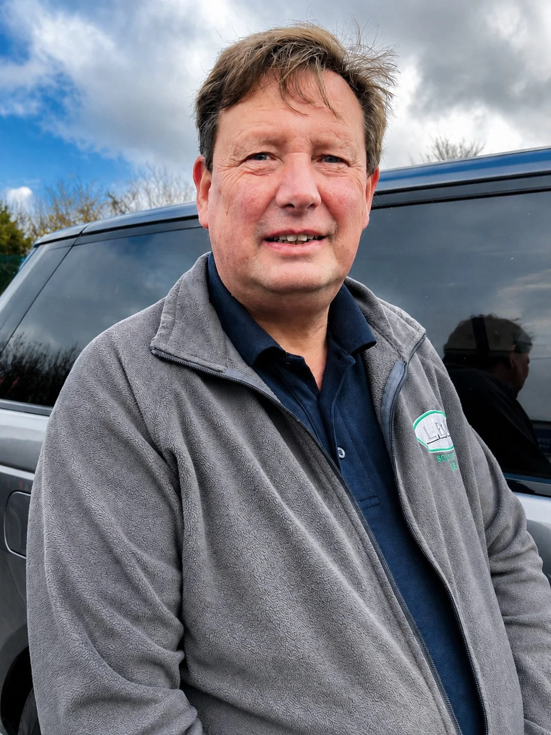 A middle-aged man with brown hair and blue eyes stands outdoors in front of a gray vehicle and a cloudy sky. He is wearing a gray zip-up jacket over a dark blue polo shirt with a logo on the chest.