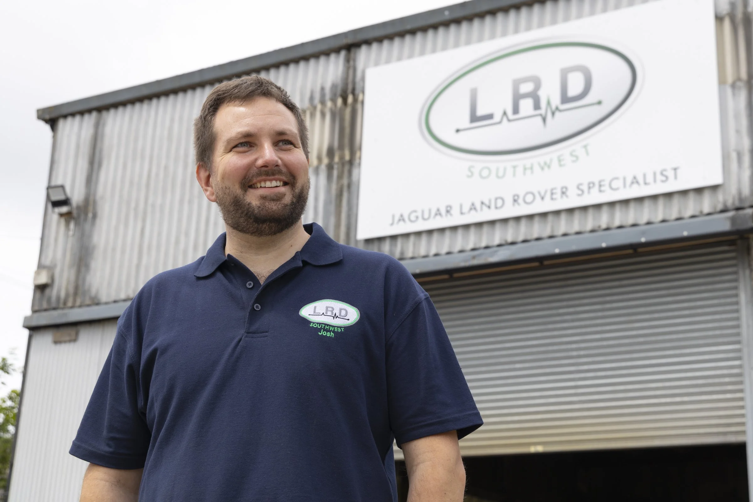 A smiling man wearing a navy polo shirt with a green and white logo, standing in front of a building with a large sign that reads 'LRD Southwest Jaguar Land Rover Specialist'.