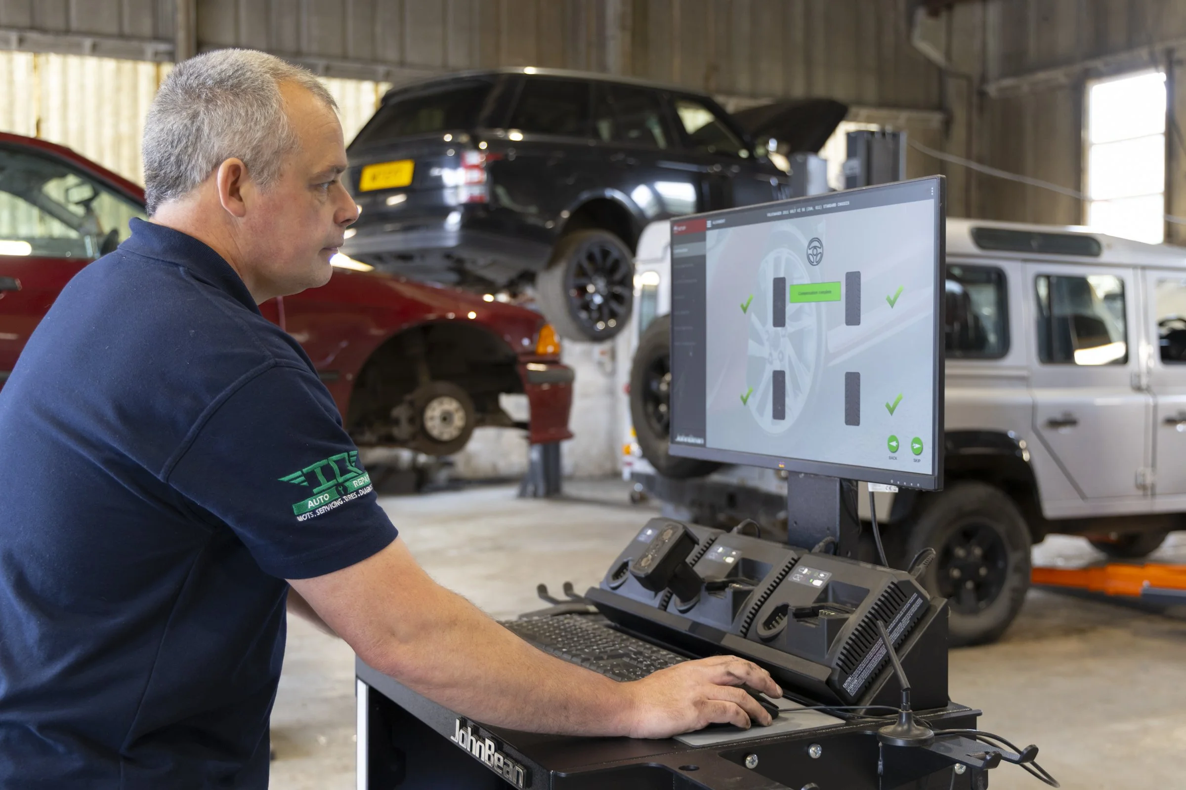 A man in a mechanic's uniform working at a computer station inside an auto repair shop, with cars on lifts in the background.