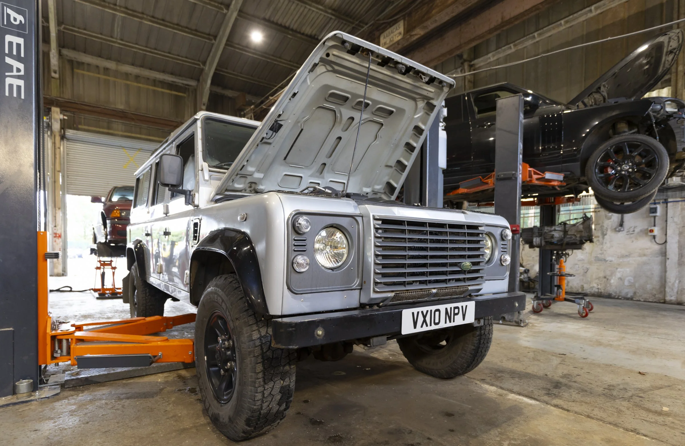 A white Land Rover Defender with its hood open, inside an auto repair shop. A black car is elevated on a hydraulic lift in the background, and tools and equipment are visible around the workshop.