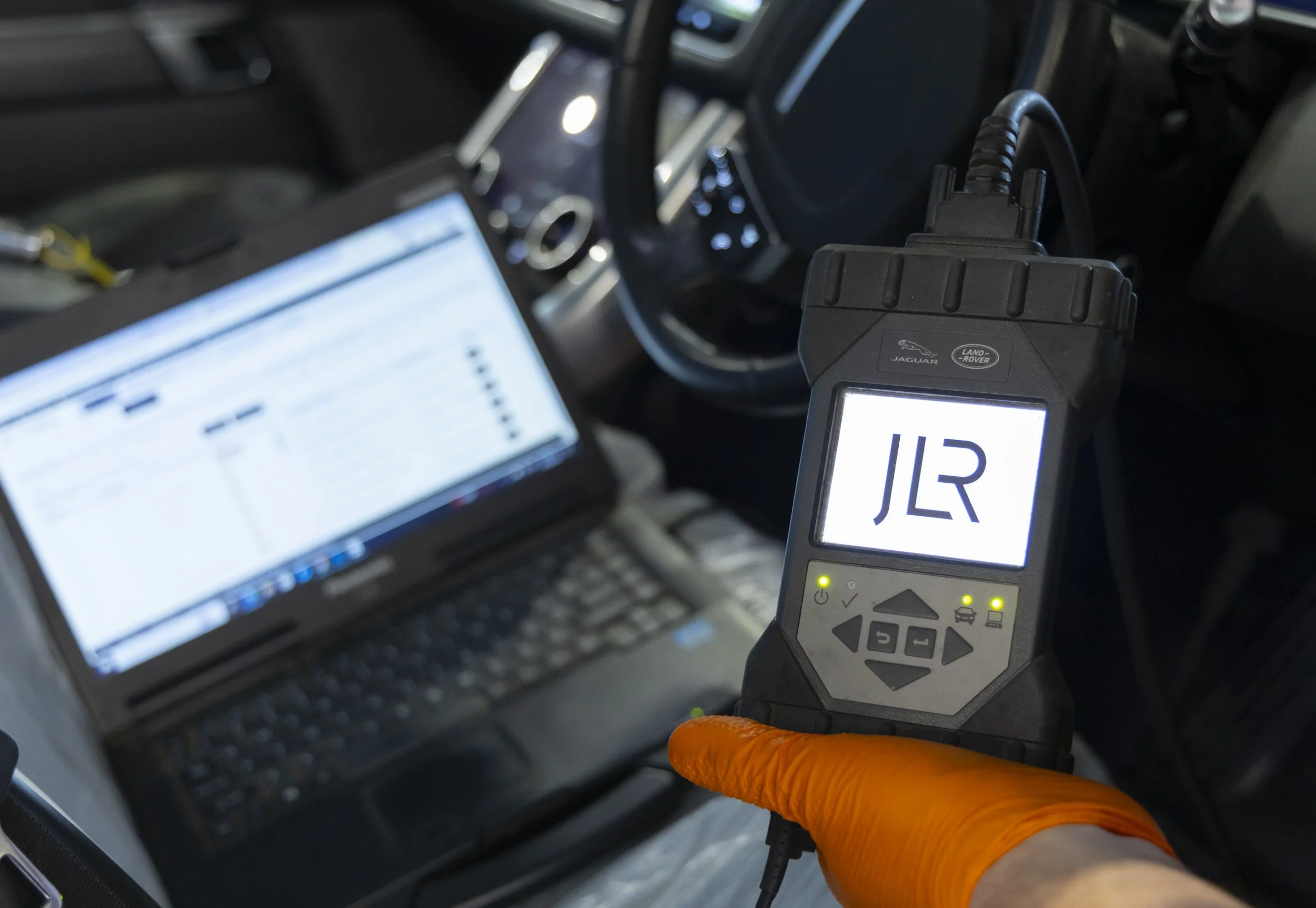 Close-up of a hand wearing an orange glove holding a diagnostic scanner connected to a car's diagnostic port; the scanner displays the code 'U3' and has buttons for operation. In the background, a laptop and part of the car's dashboard are visible.