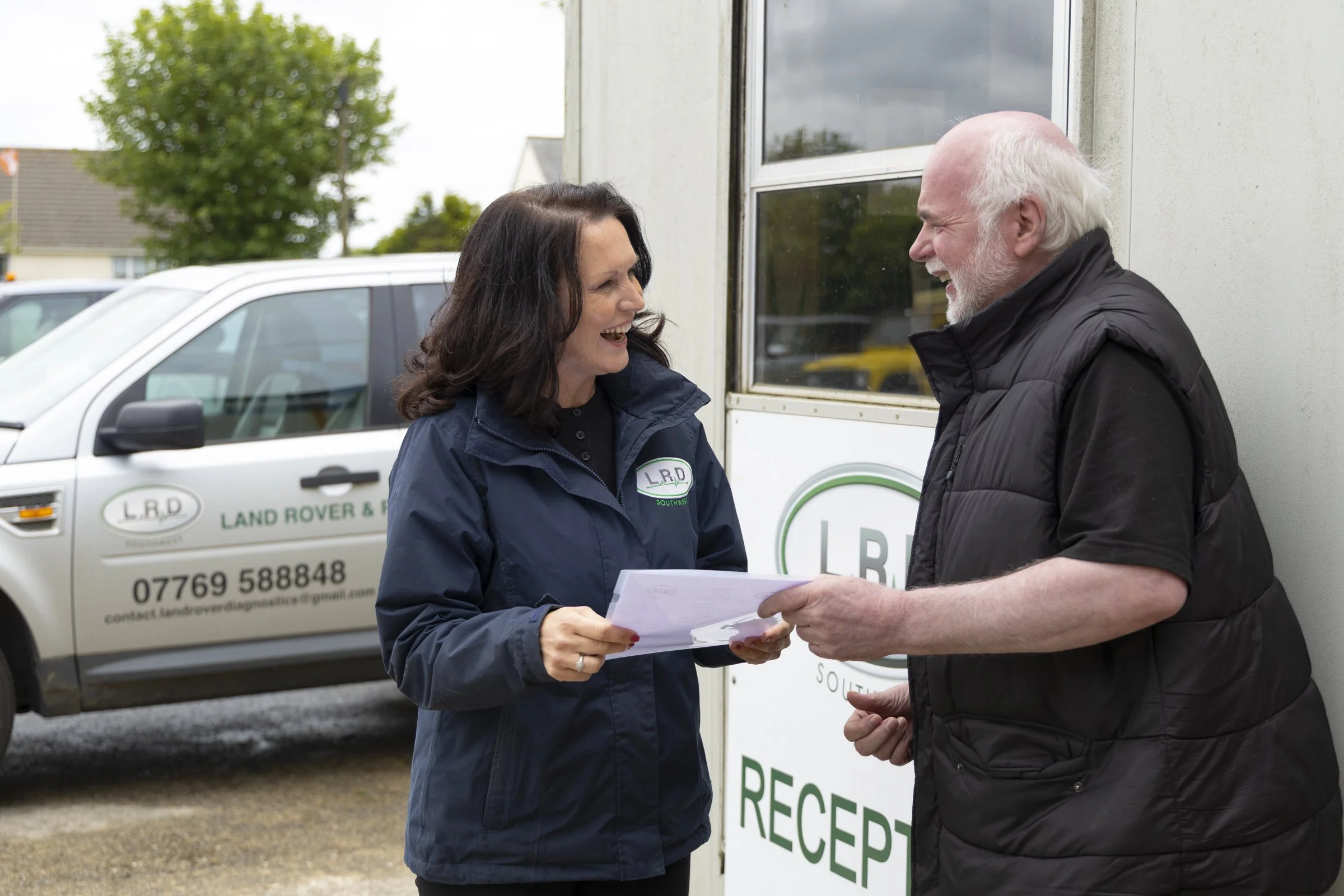 A woman and a man are standing outside, smiling and talking. The woman is holding some papers while the man is holding a small item. In the background, there is a white vehicle with a logo that reads 'LRD' and a phone number. The woman is wearing a navy blue jacket, and the man is wearing a black vest. They are near a sign that says 'RECE'.