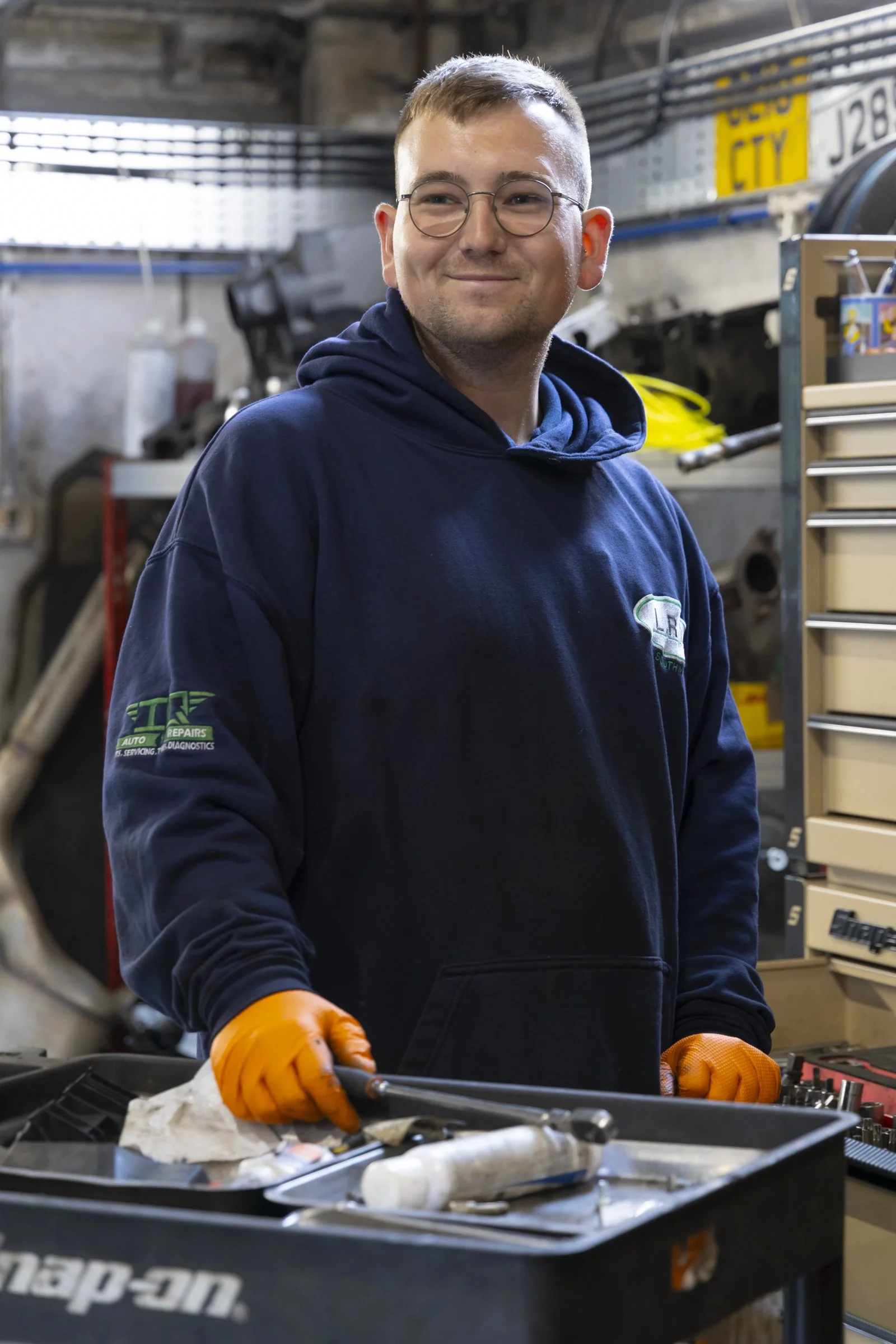 A young man with short brown hair, glasses, and a slight smile, wearing an ID badge and orange gloves, working on car maintenance in a garage.