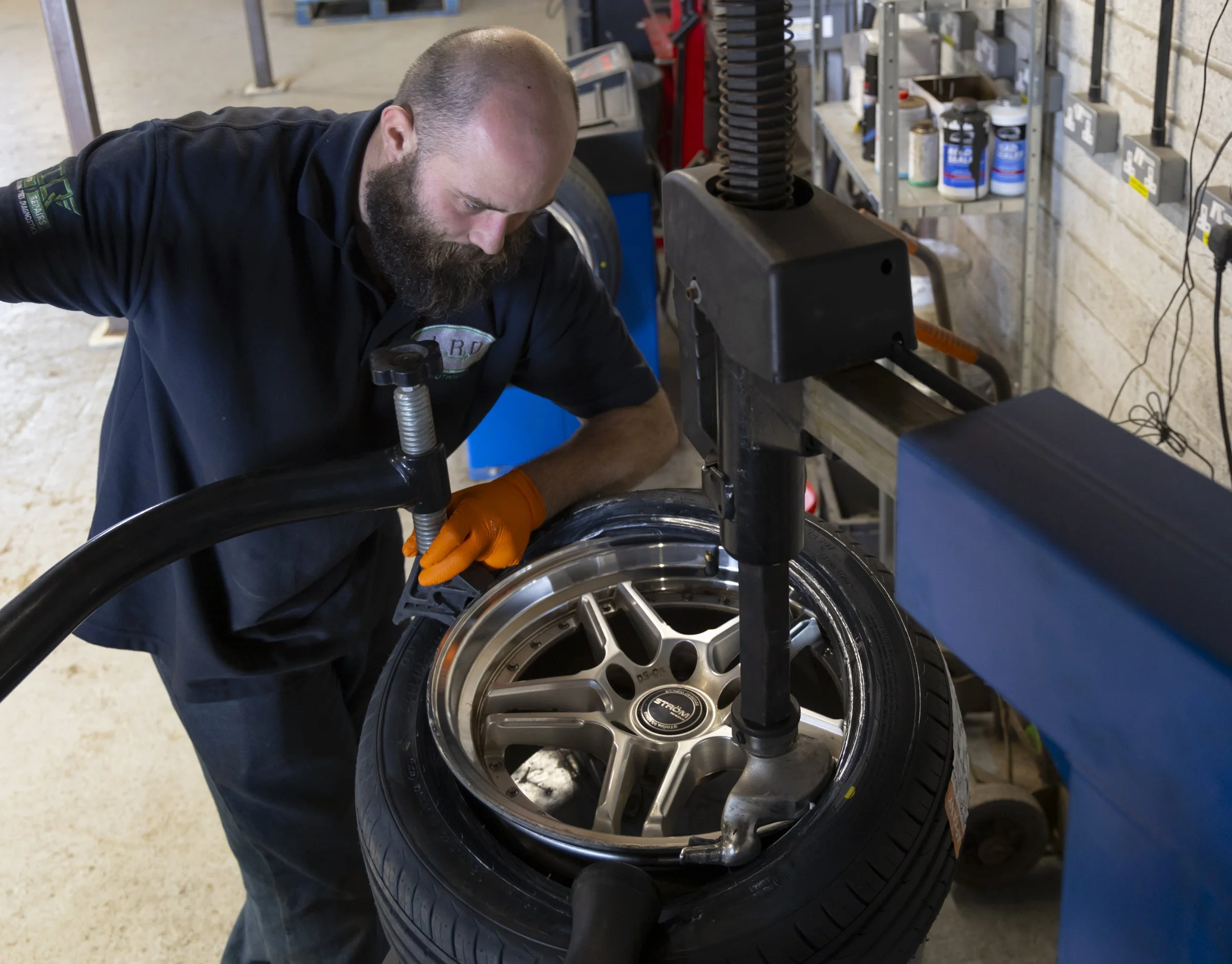 A man working on a car wheel in a garage workshop, wearing an orange glove and concentrating on the tire fitting process.