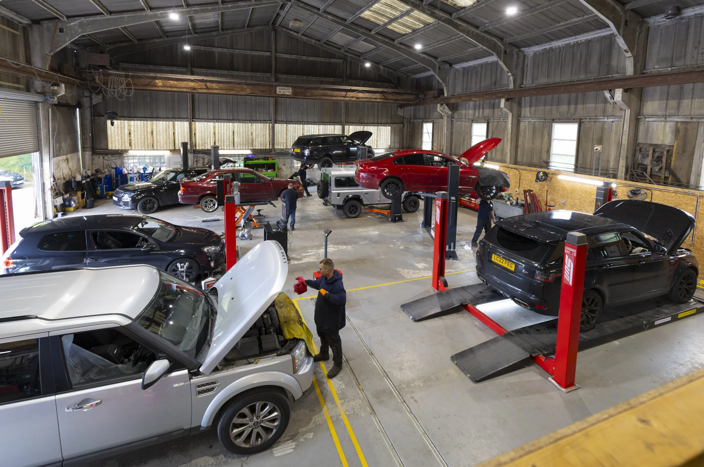 Inside a spacious auto repair garage with multiple cars being serviced, including a silver SUV with its hood open, several black and red cars on lifts, and mechanics working on vehicles. The garage has a high, metal ceiling and various tools and equipment along the walls.