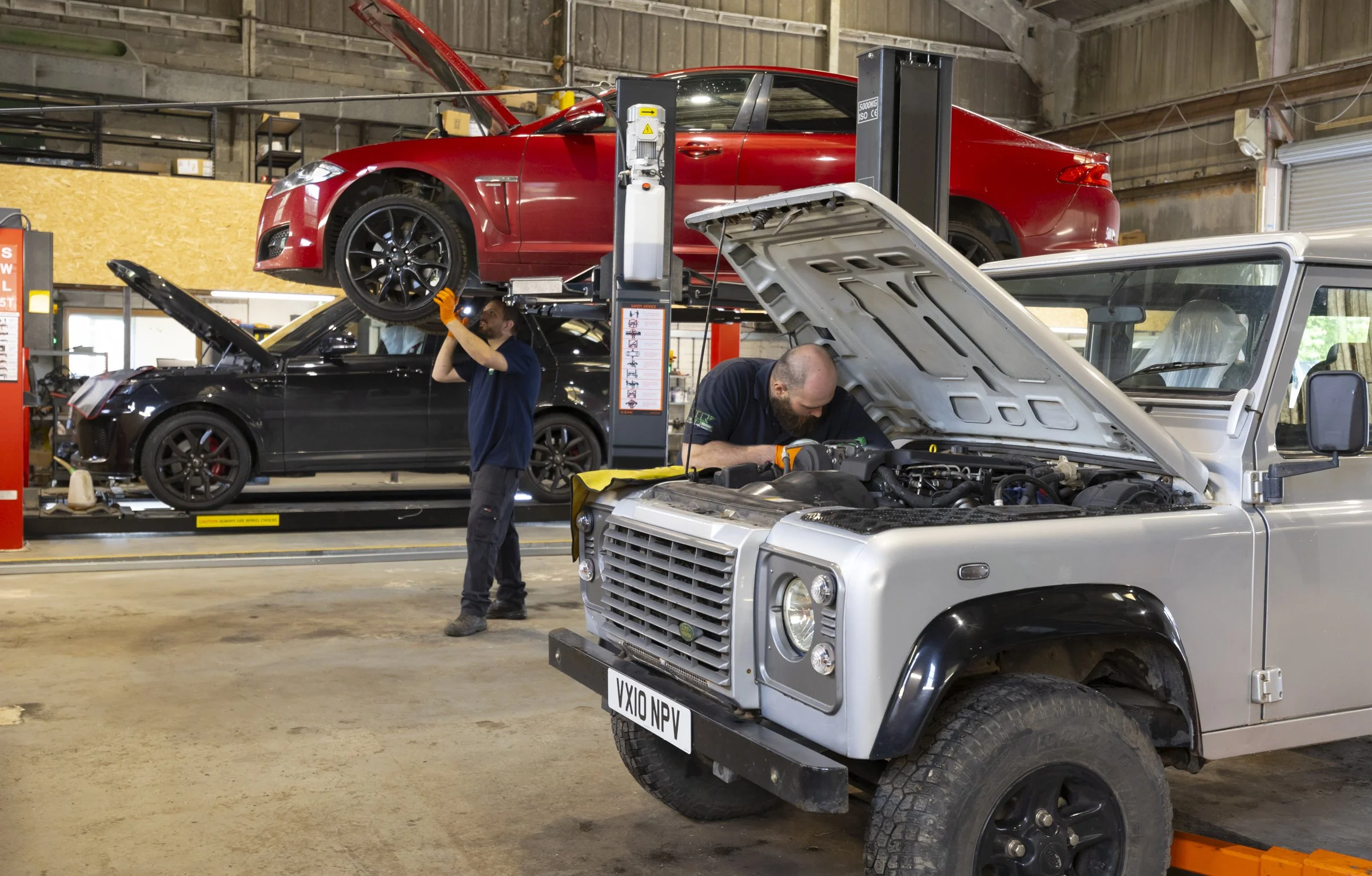 Inside an automotive repair shop with a red car on a lift, two mechanics working, and two additional cars with open hoods being serviced.