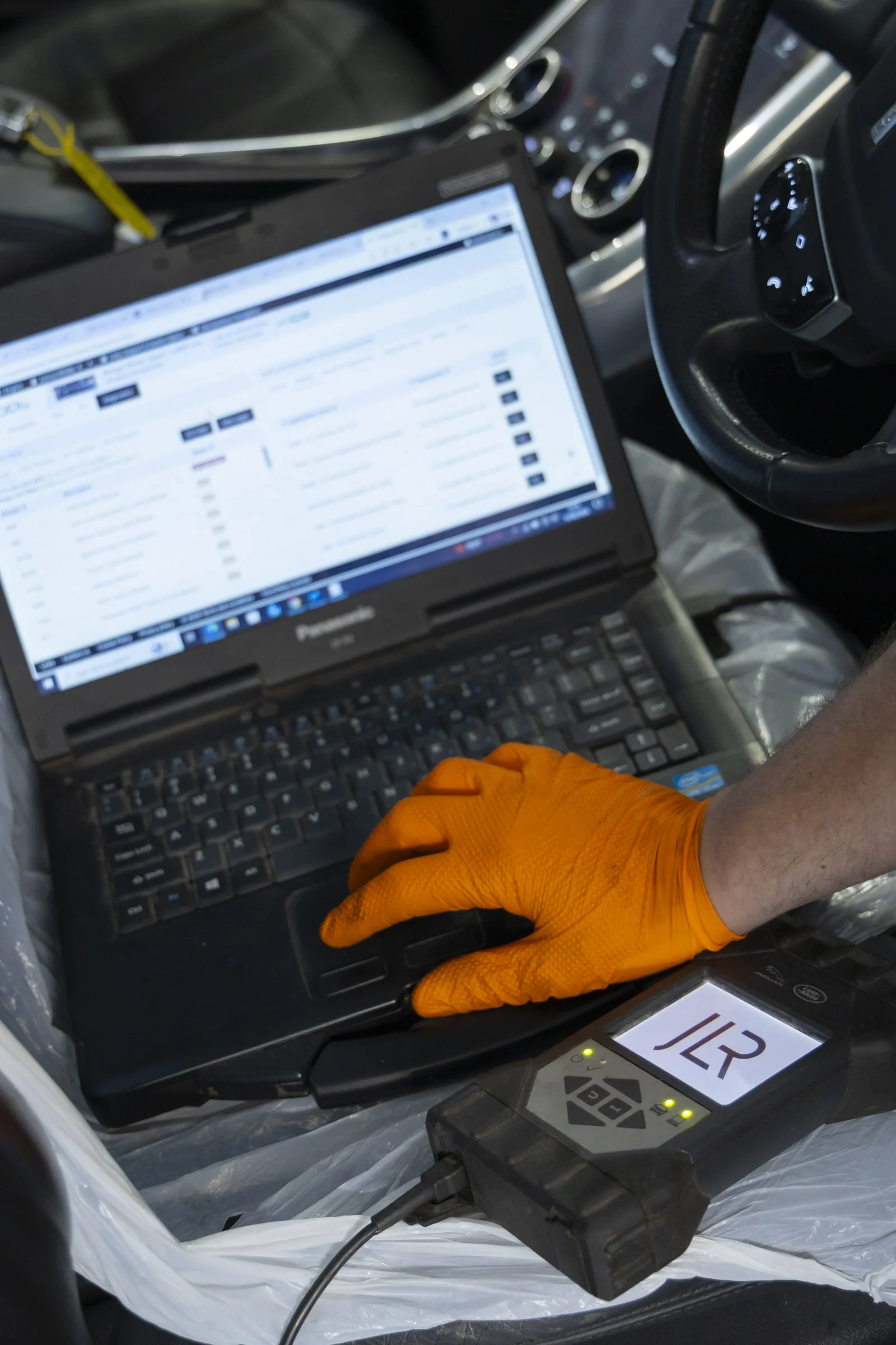Inside a vehicle, a person wearing orange gloves is working with a laptop and electronic devices, possibly conducting diagnostic or technical work.