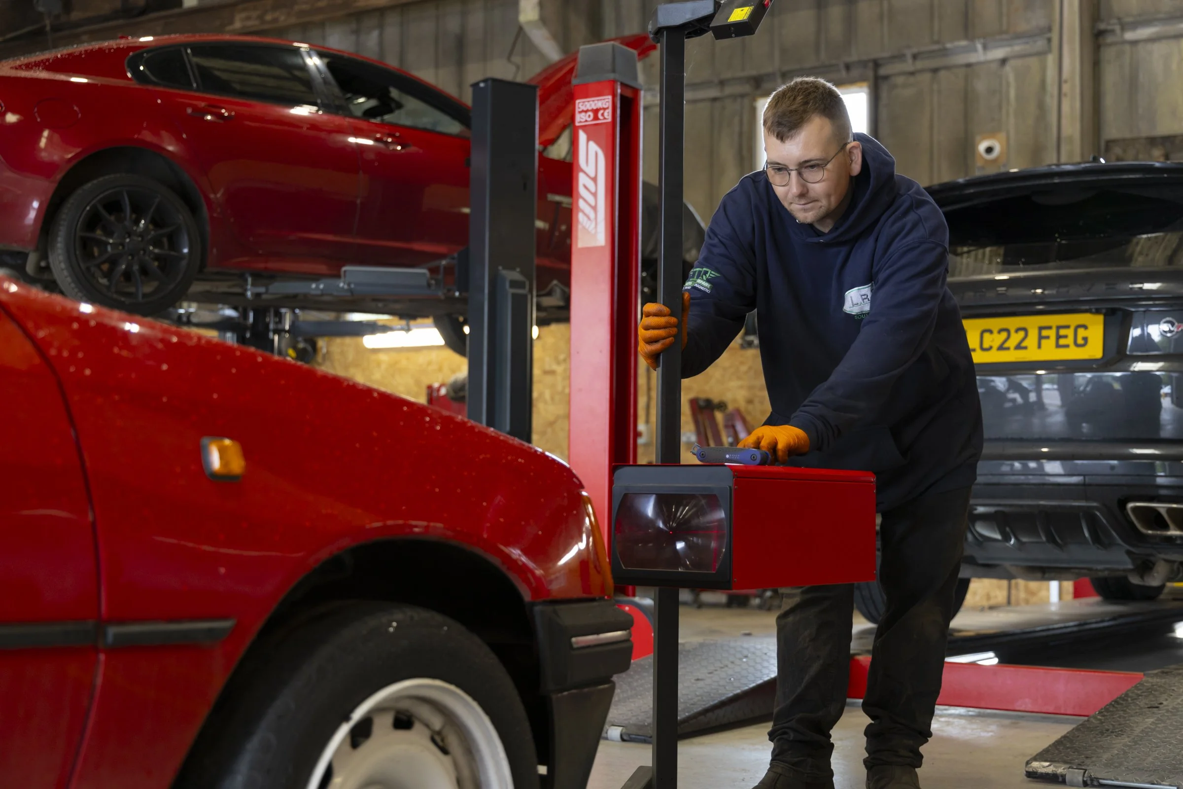 A man wearing glasses and orange gloves working on a car in an auto repair shop, with a red sports car elevated on a lift in the background and another vehicle visible.