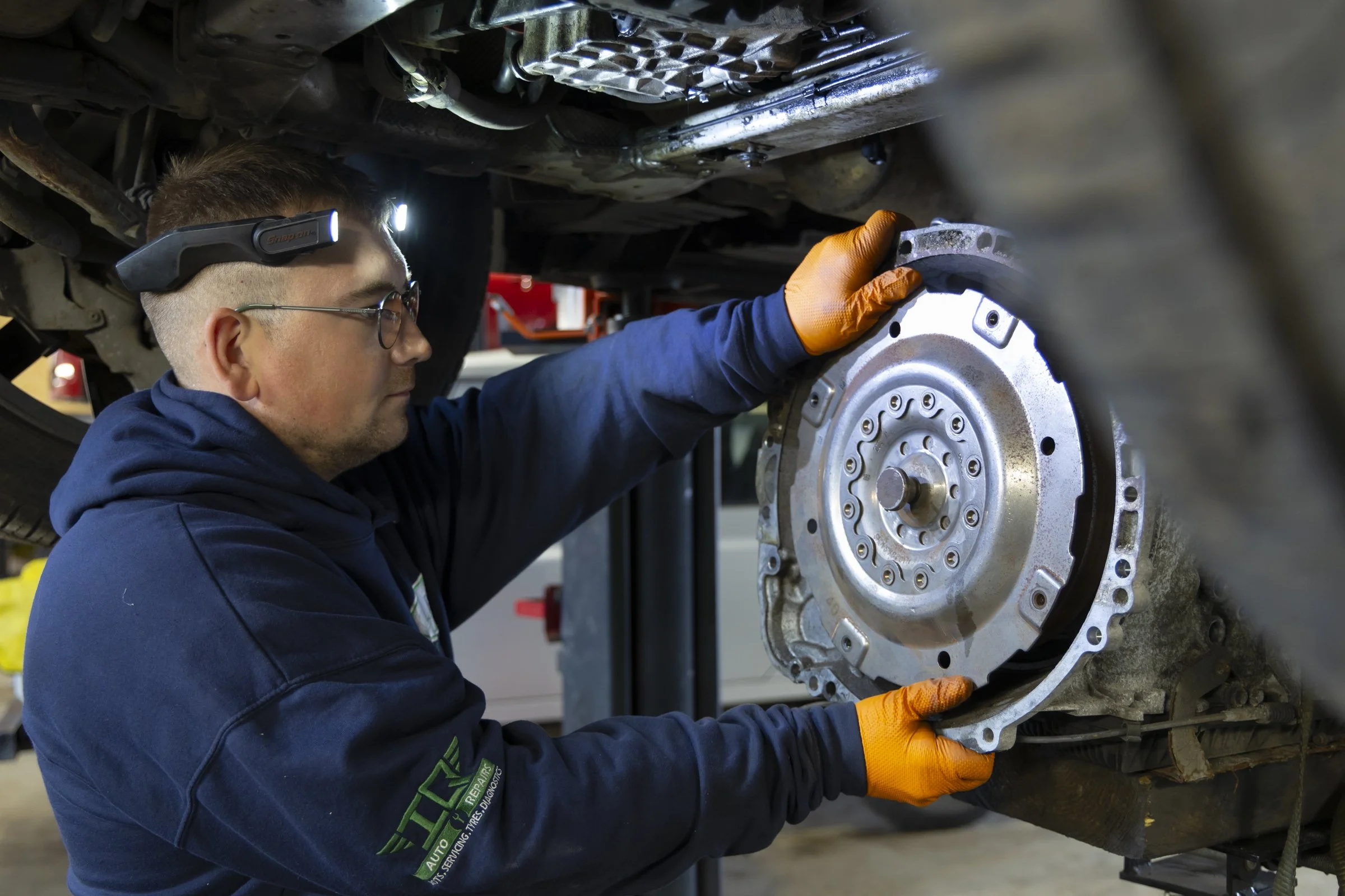 A mechanic wearing safety goggles and orange gloves working under a vehicle, handling a large metal engine component in an auto repair shop.