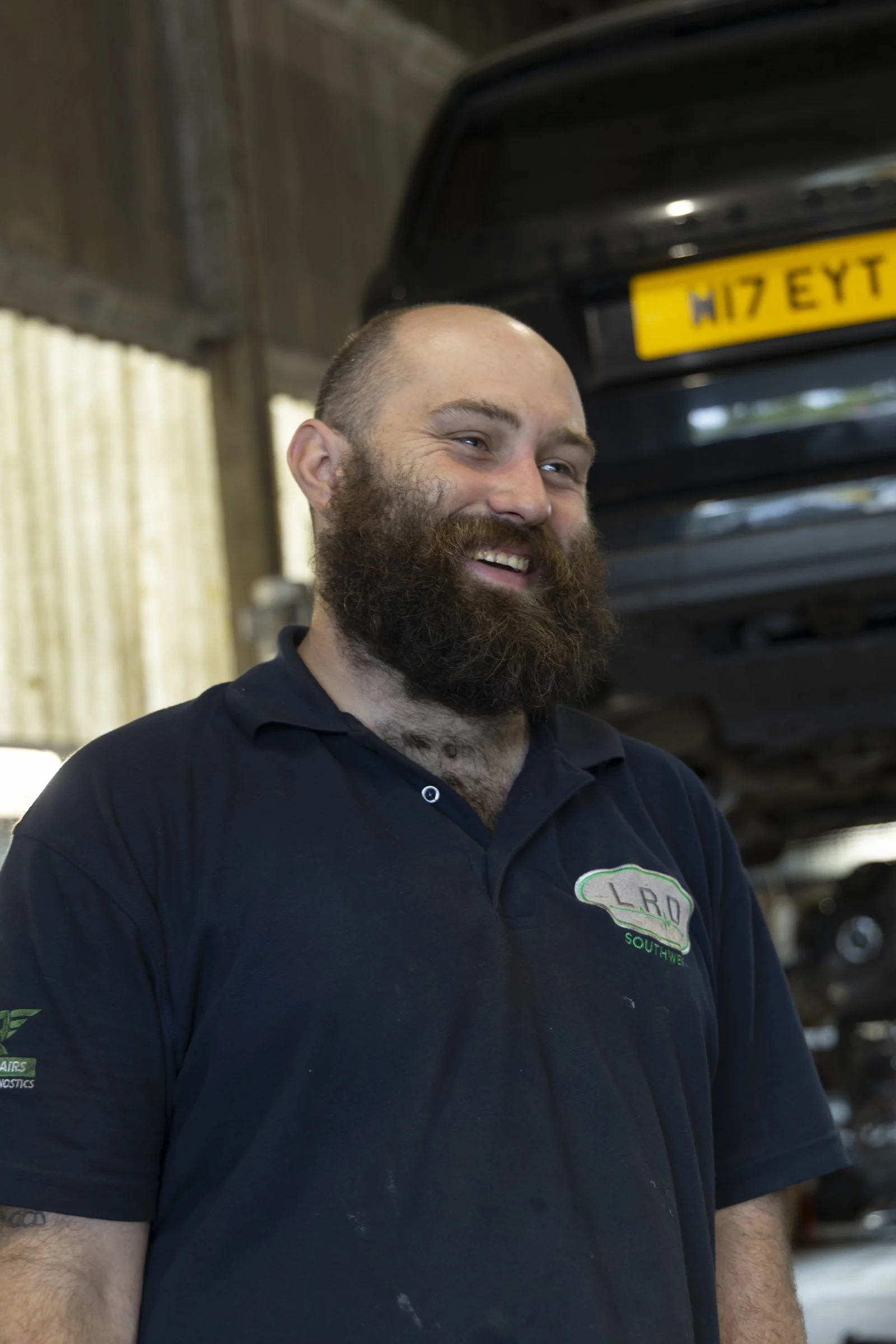 A smiling man with a beard wearing a black polo shirt with a logo, standing inside a workshop or garage with blurry tools and vehicle parts in the background.