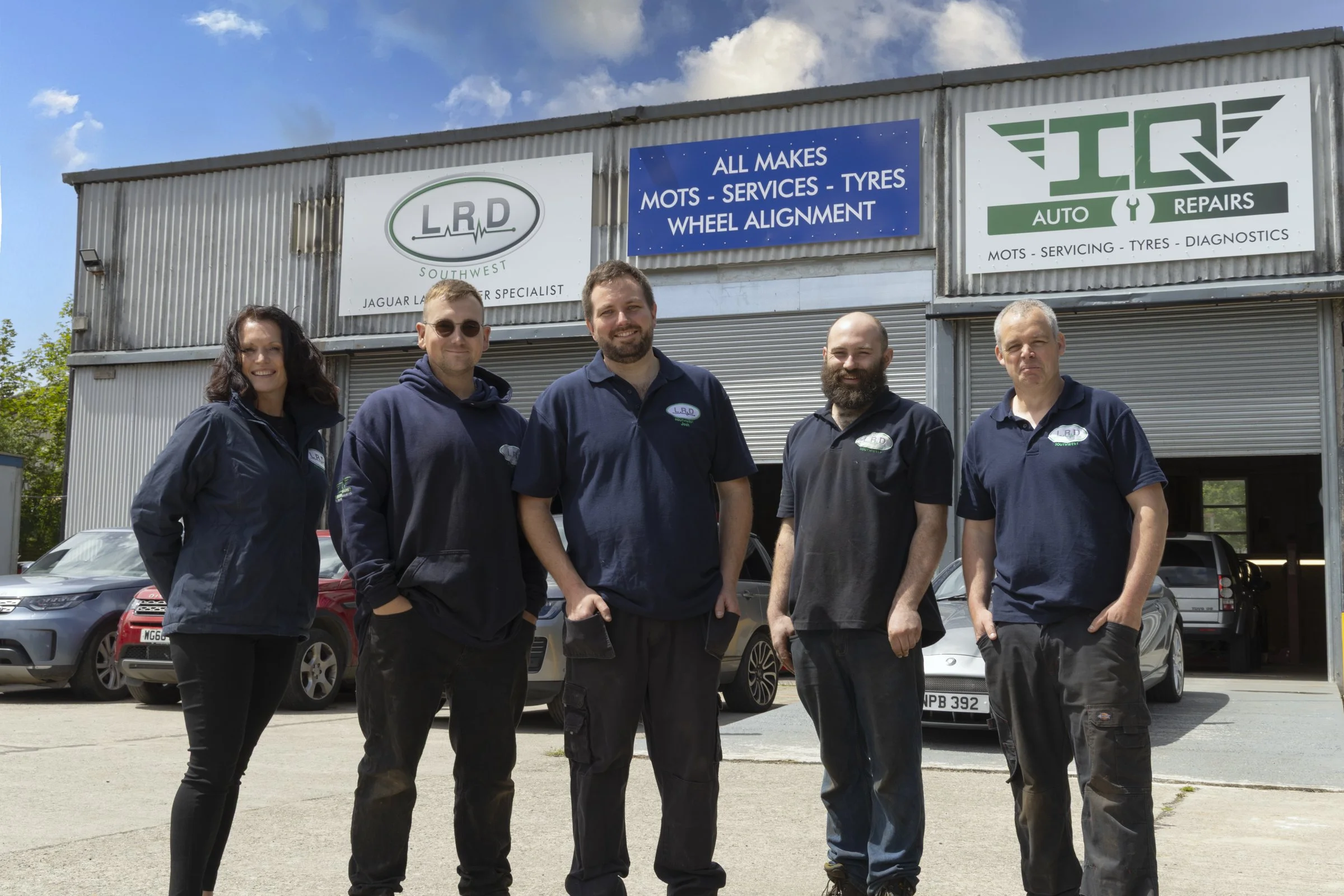 Group of five auto repair technicians standing in front of an auto repair shop with signs listing services like MOTs, servicing, tires, and wheel alignment. The technicians are smiling and dressed in dark uniforms.