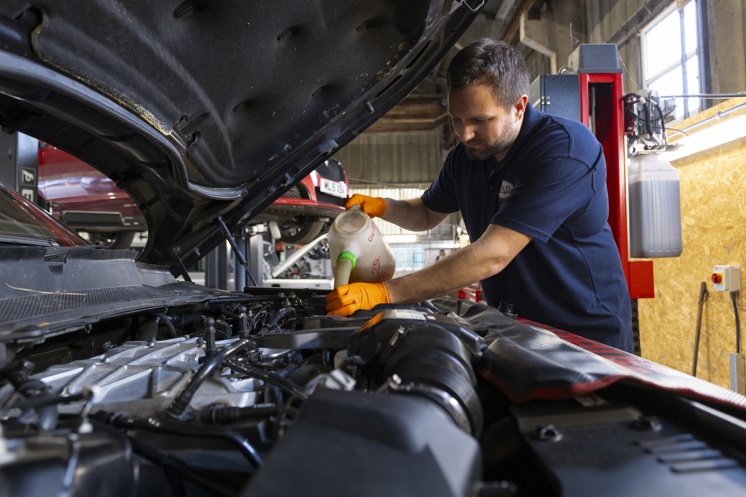 A mechanic working under the hood of a car in a garage, pouring fluid into the engine.