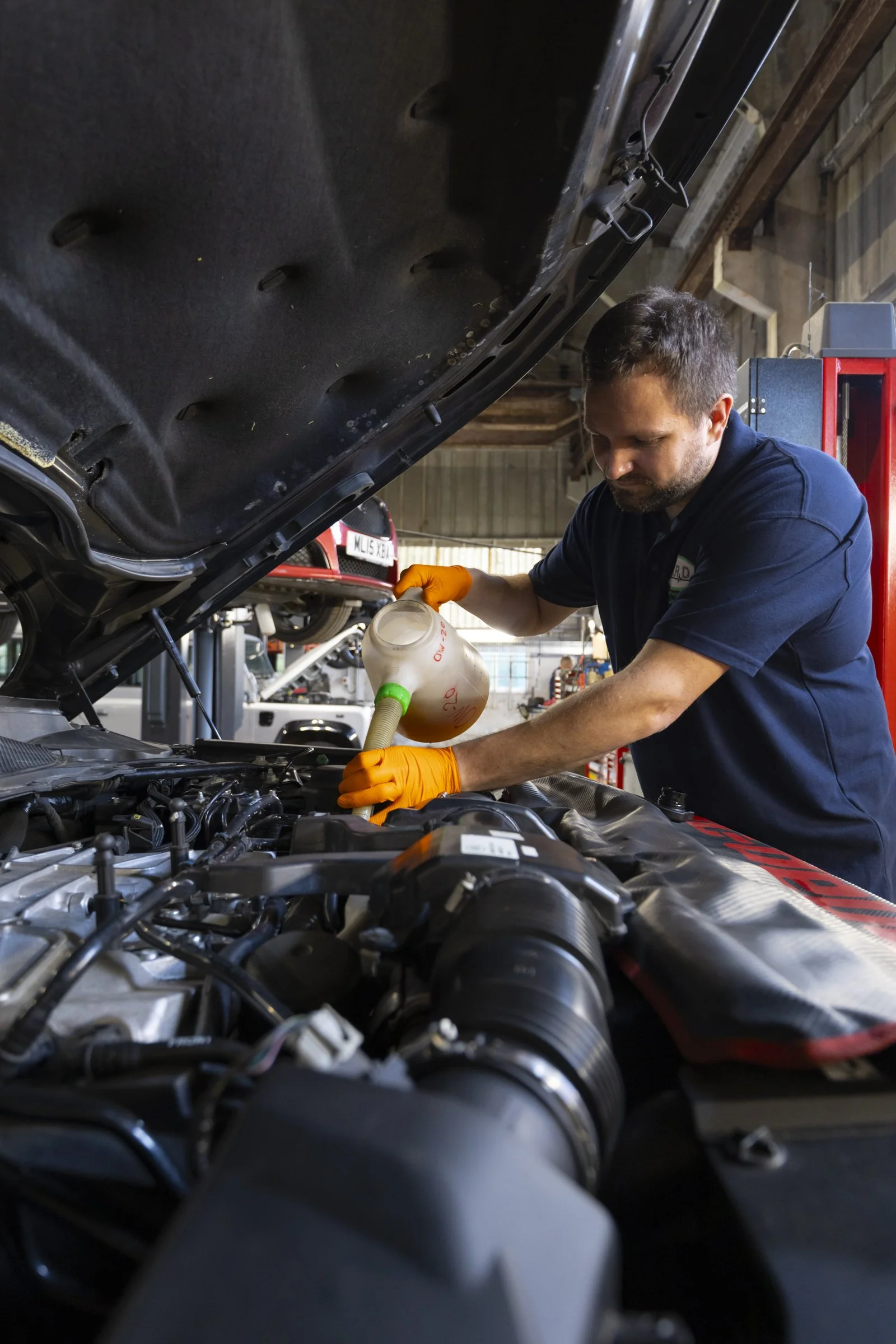 A mechanic wearing orange gloves adds oil to a car engine with the hood open inside a garage.