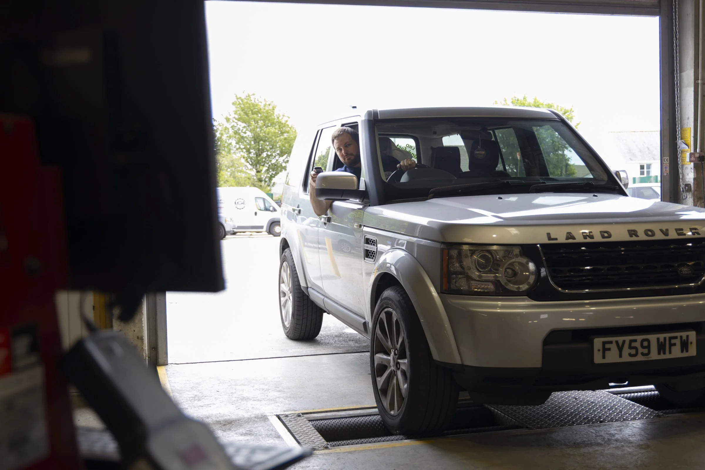 A man with a beard and short hair looking out from the passenger side of a silver Land Rover vehicle, parked inside a garage. The garage door is open, revealing trees and another vehicle outside.