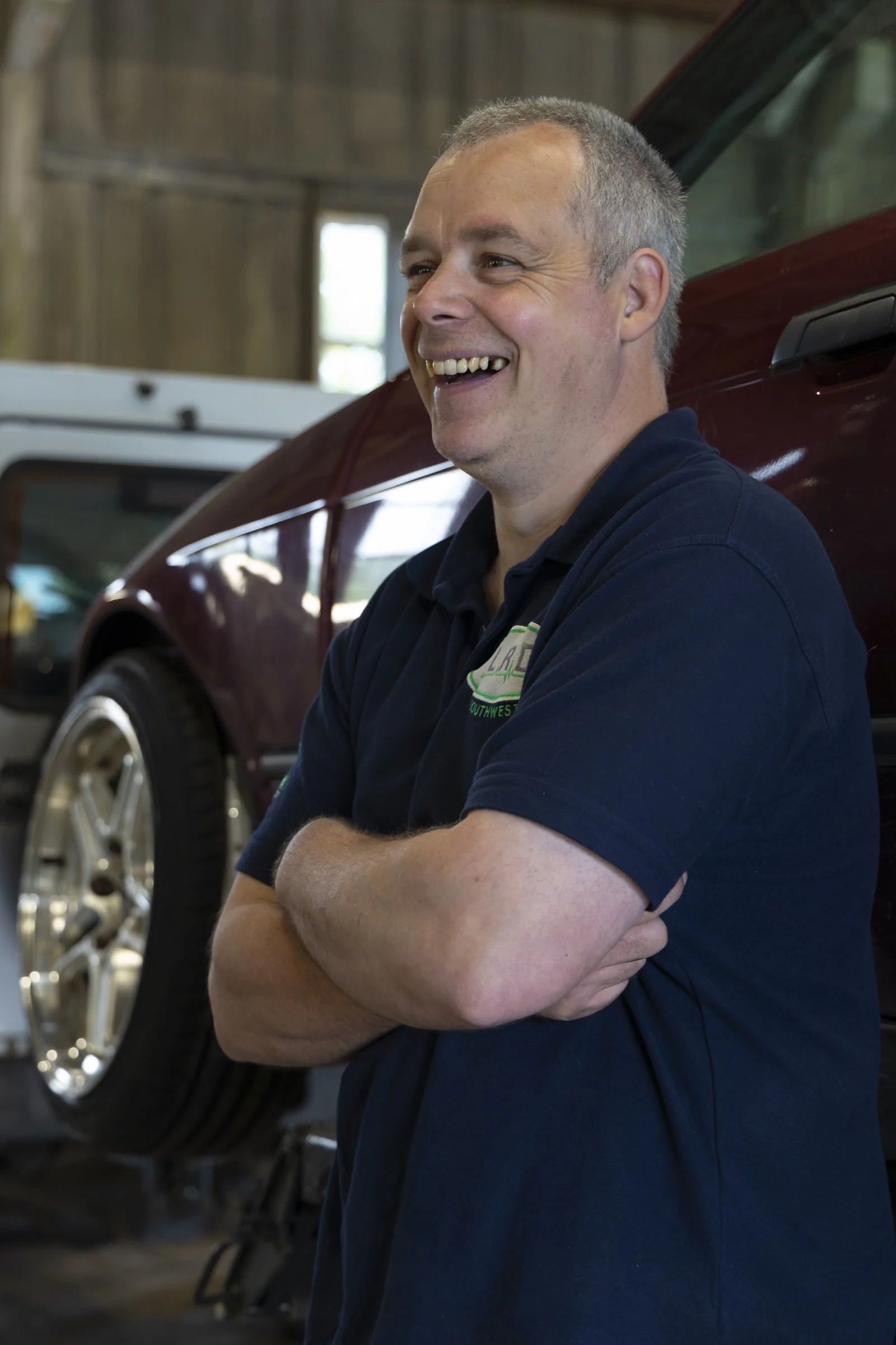 A man with short, gray hair smiling with his arms crossed, standing next to a red vehicle inside a wooden building with a window in the background.