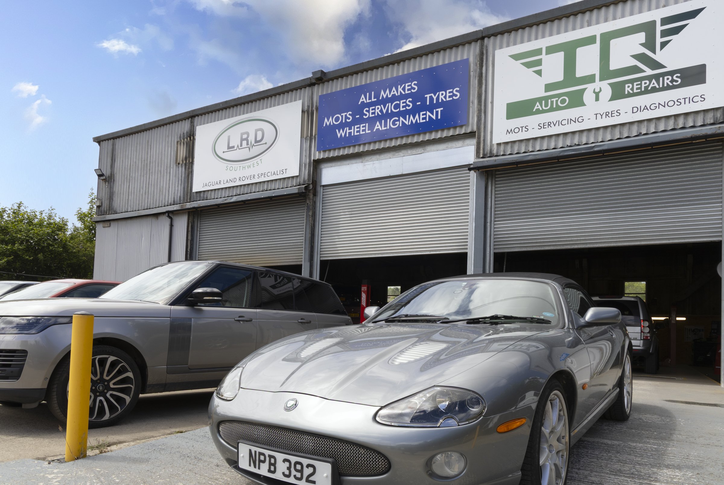 Image of a car repair shop with signs for auto services, tire services, wheel alignment, diagnostics, and brand logos, with several parked cars in front.