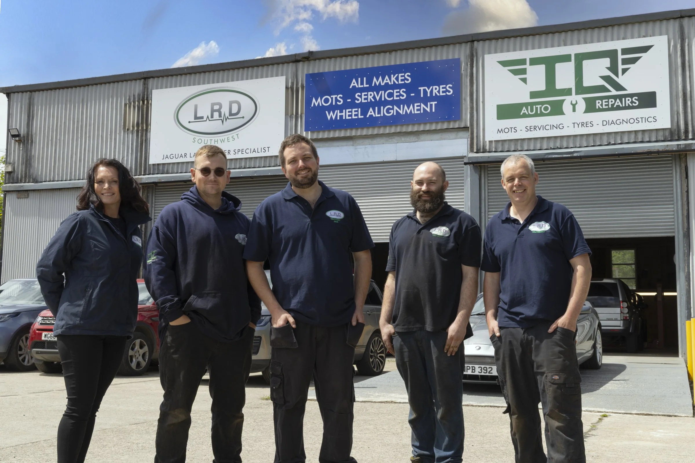 Group of five auto repair technicians standing in front of an auto shop with signs for auto services, tire services, and wheel alignment. They are smiling and dressed in work uniforms.