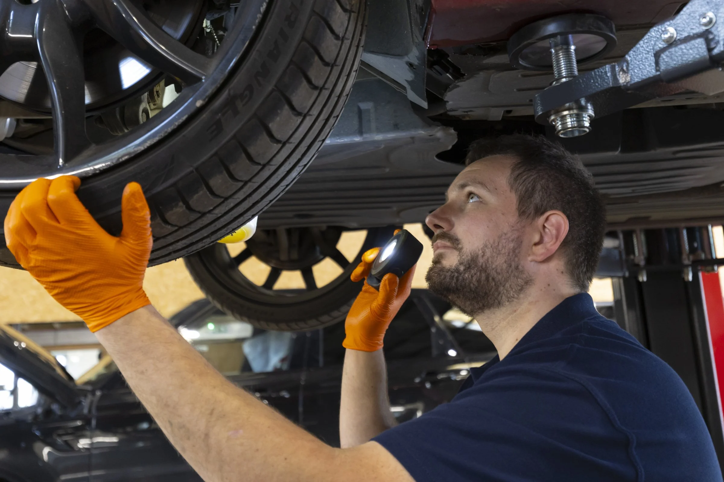 A mechanic working underneath a car, inspecting or repairing the vehicle while holding a flashlight and wearing orange gloves.