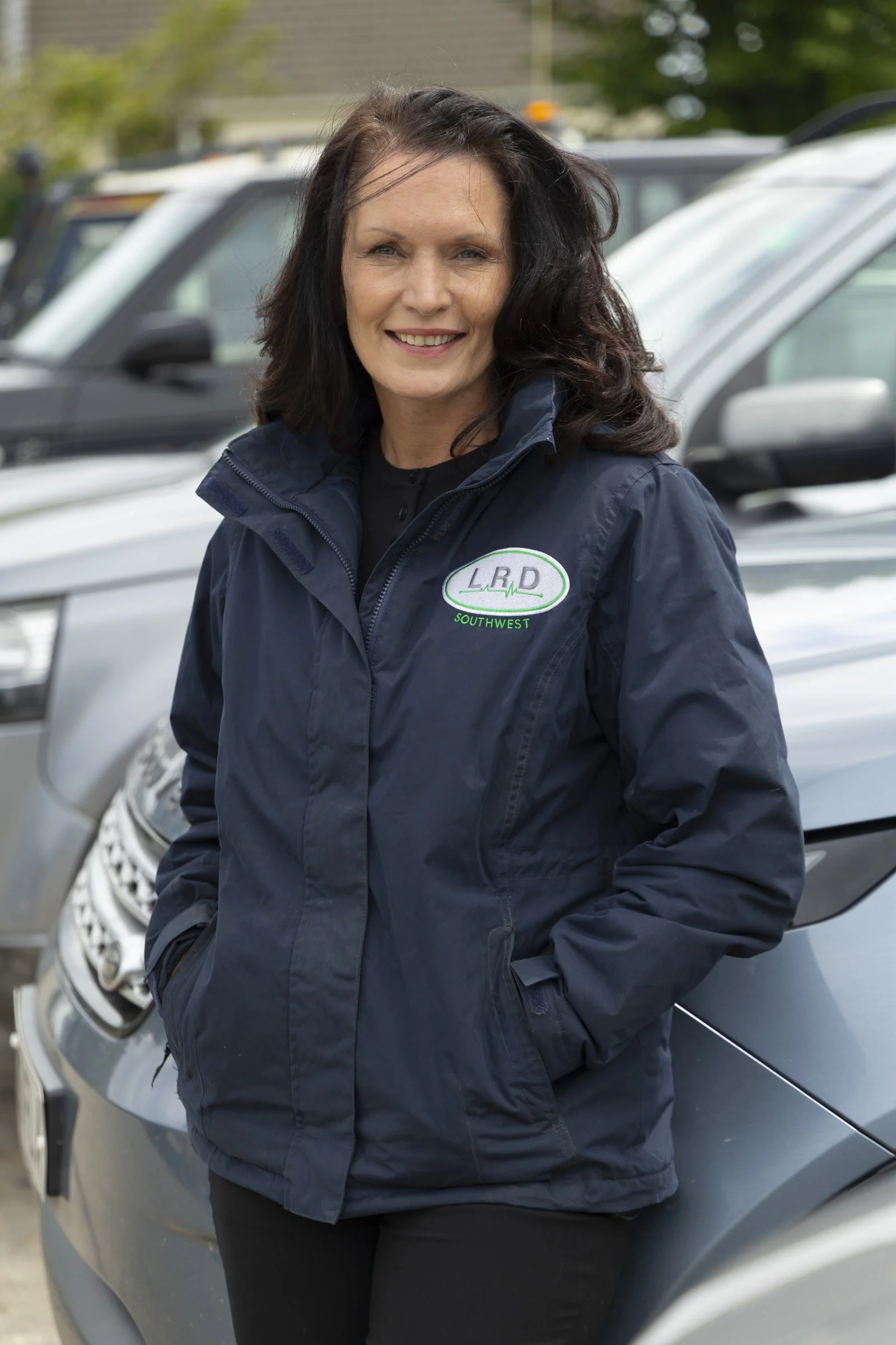 A woman with dark hair smiling, standing in front of parked cars, wearing a navy blue jacket with a logo that says 'L.R.D SW Southwest'.