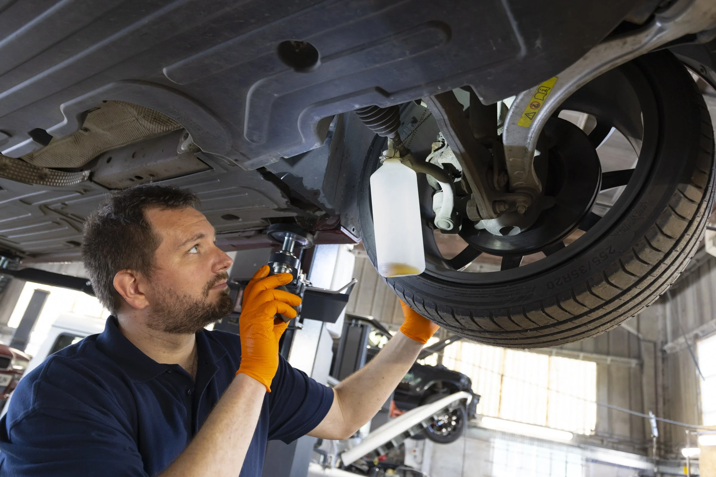 Man working under a raised car, inspecting or repairing the undercarriage, wearing orange gloves, inside a garage or workshop.