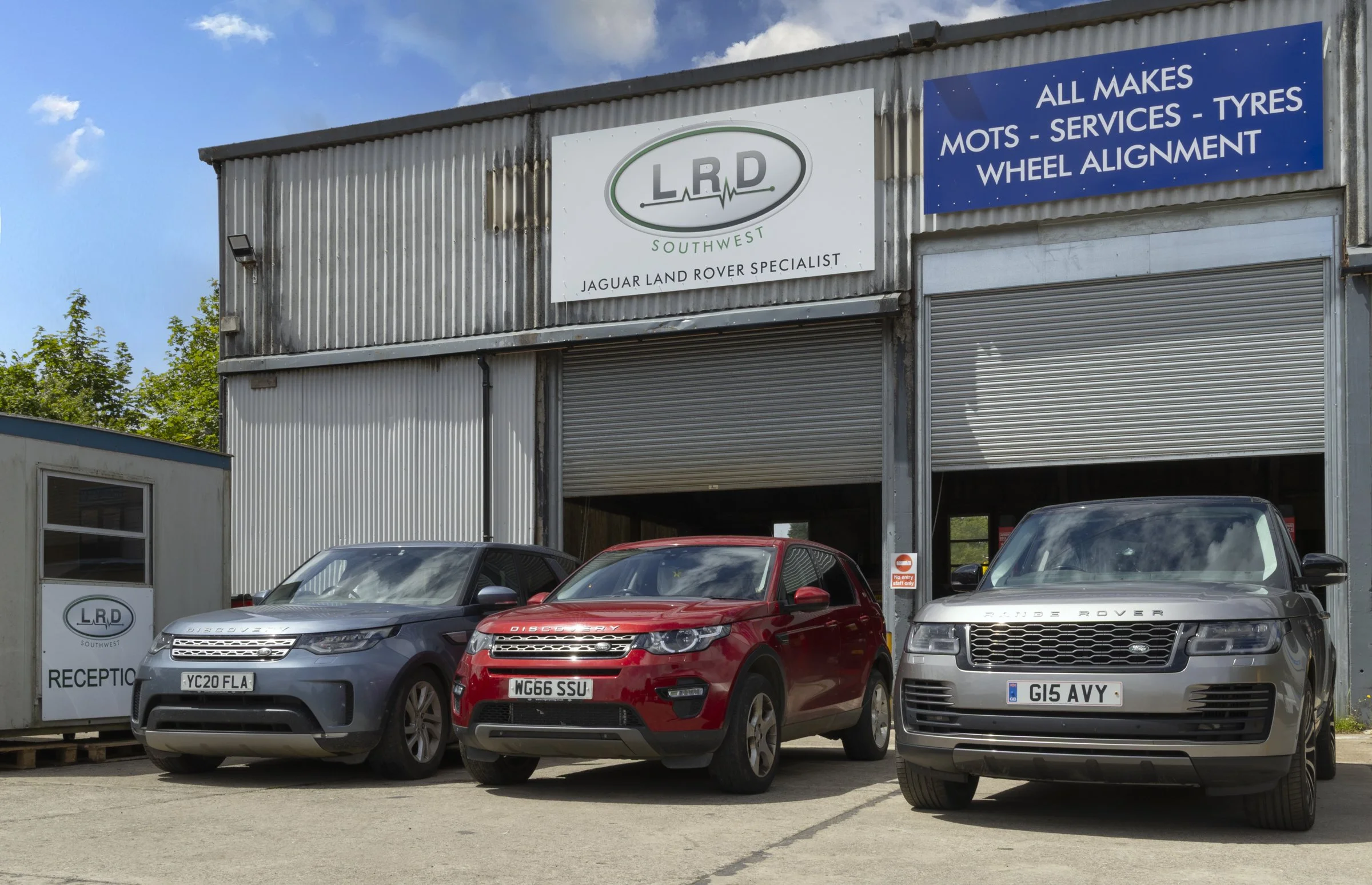 A garage with cars parked in front, featuring signs for a Land Rover specialist and vehicle services including MOTs, servicing, and wheel alignment.