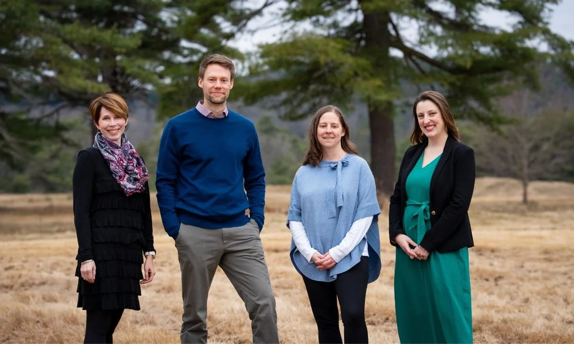 Four people standing outdoors in a field with trees in the background, smiling at the camera.
