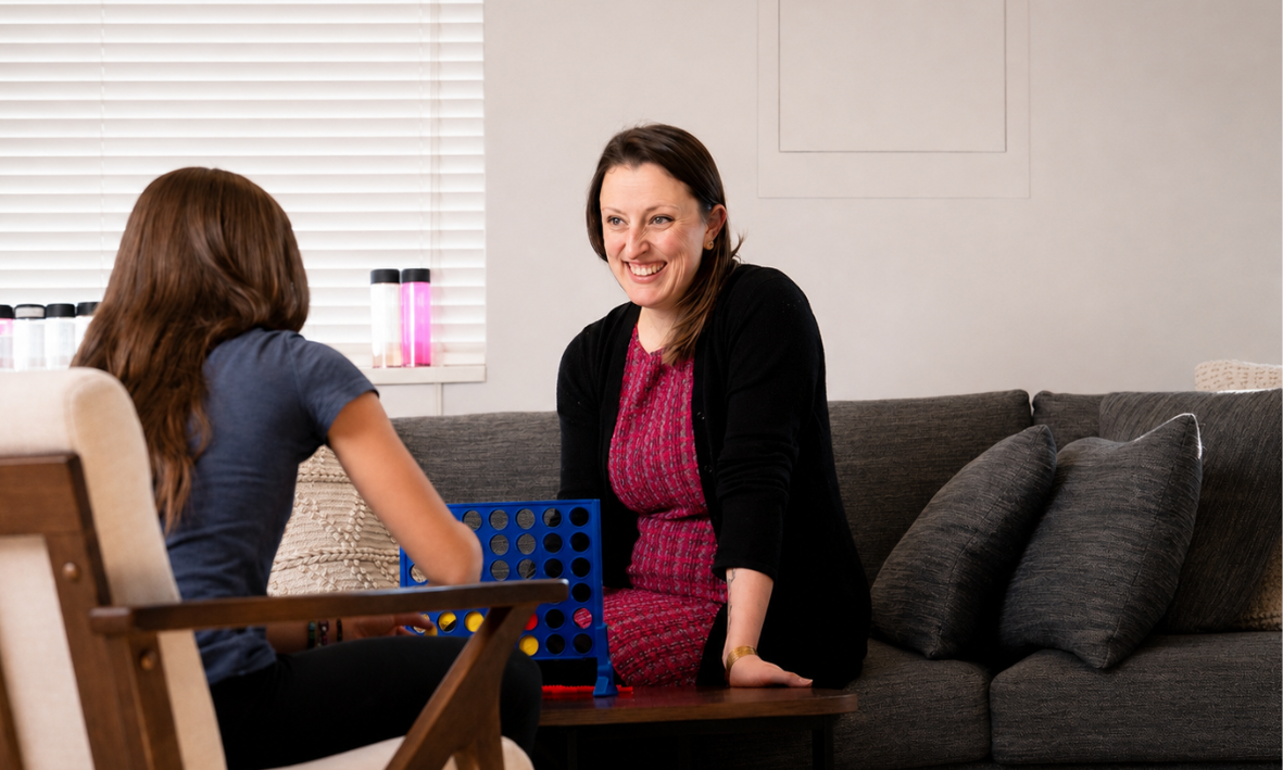 Woman and girl playing Connect Four on a couch in a bright room.