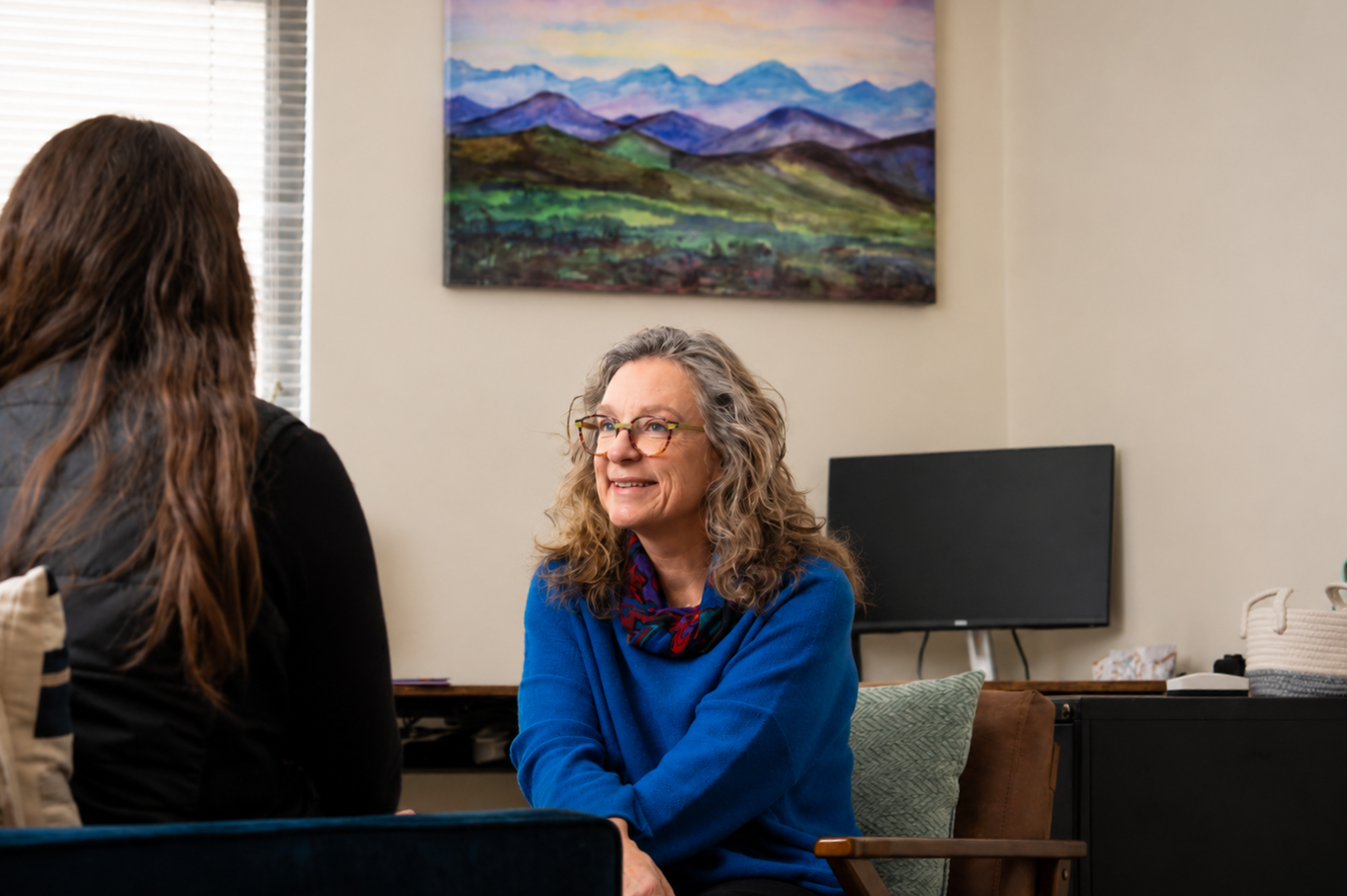 A woman with curly gray hair, glasses, and a blue sweater sitting in an office, smiling during a conversation with another woman with long brown hair.