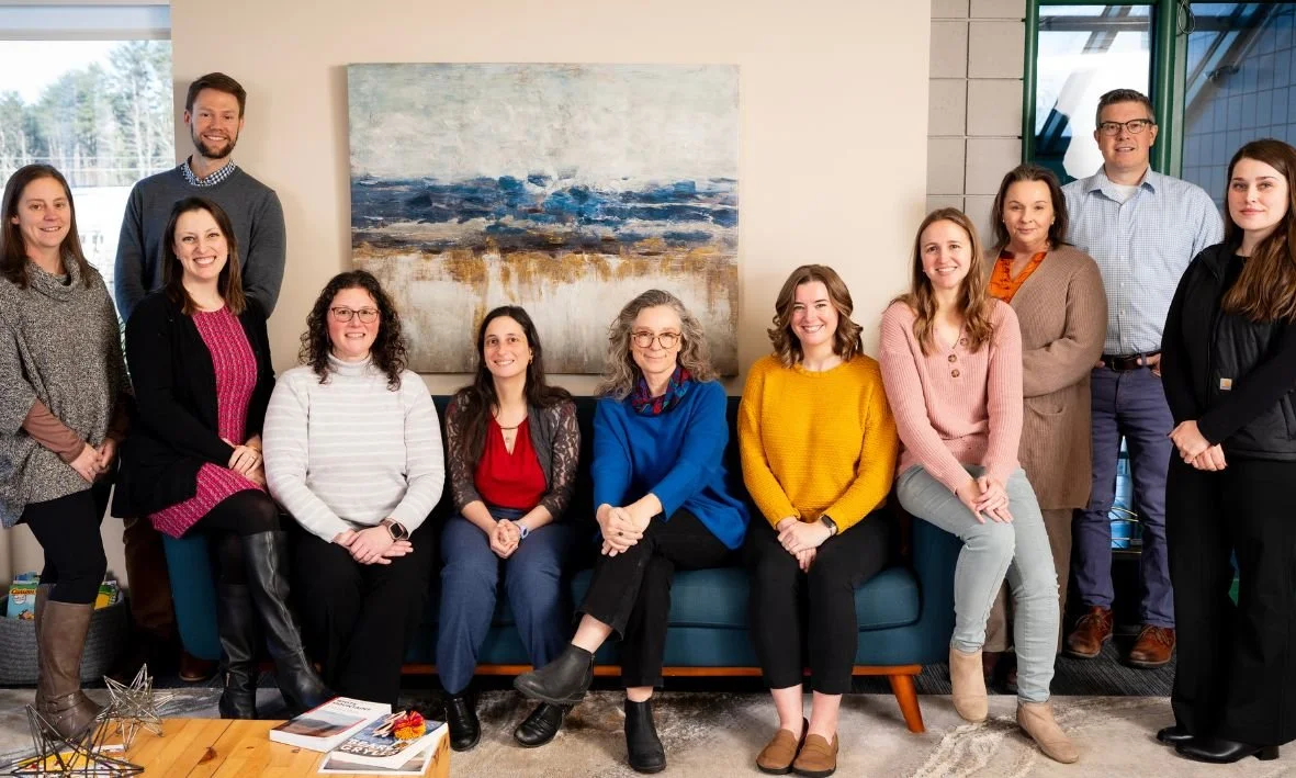 A group of twelve diverse people, including men and women, posing in a modern office with a large abstract painting behind them.