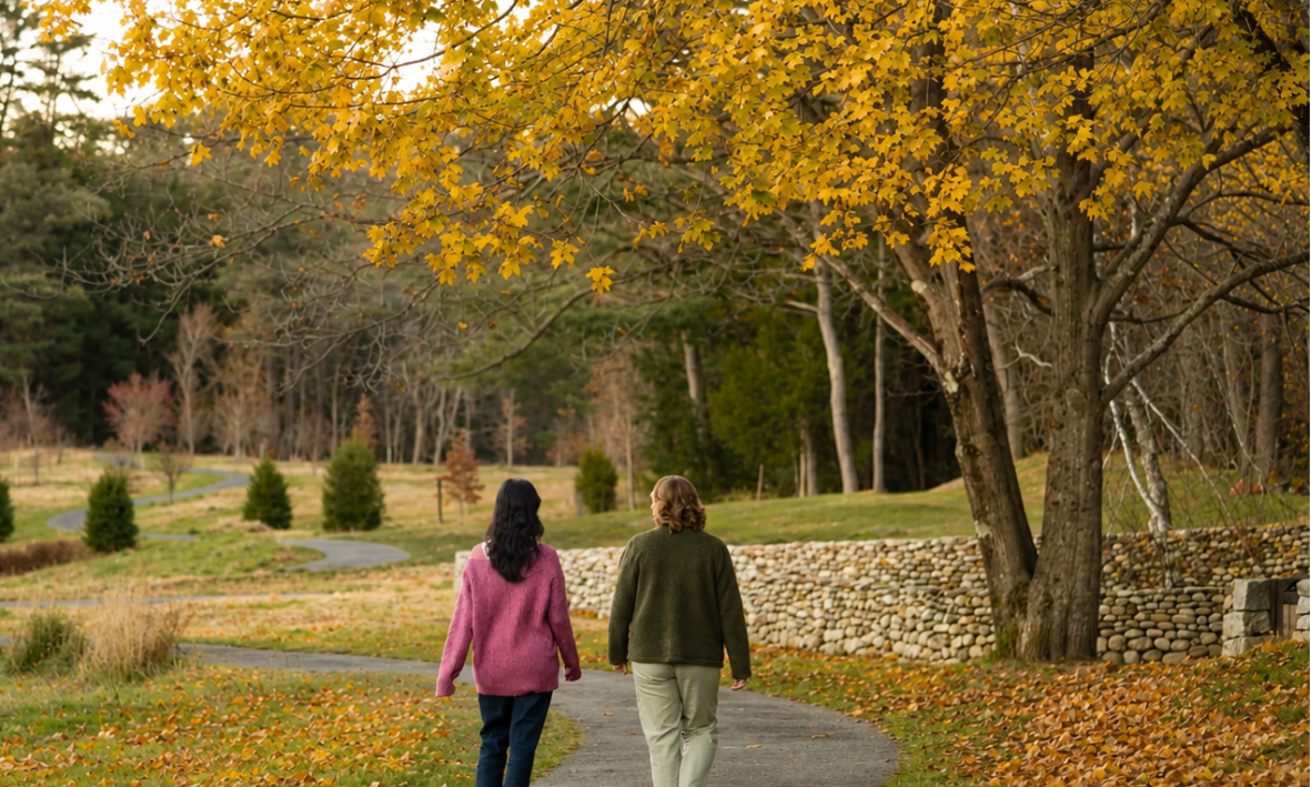 Two people walking on a winding path in a park during fall, surrounded by trees with yellow and orange leaves.